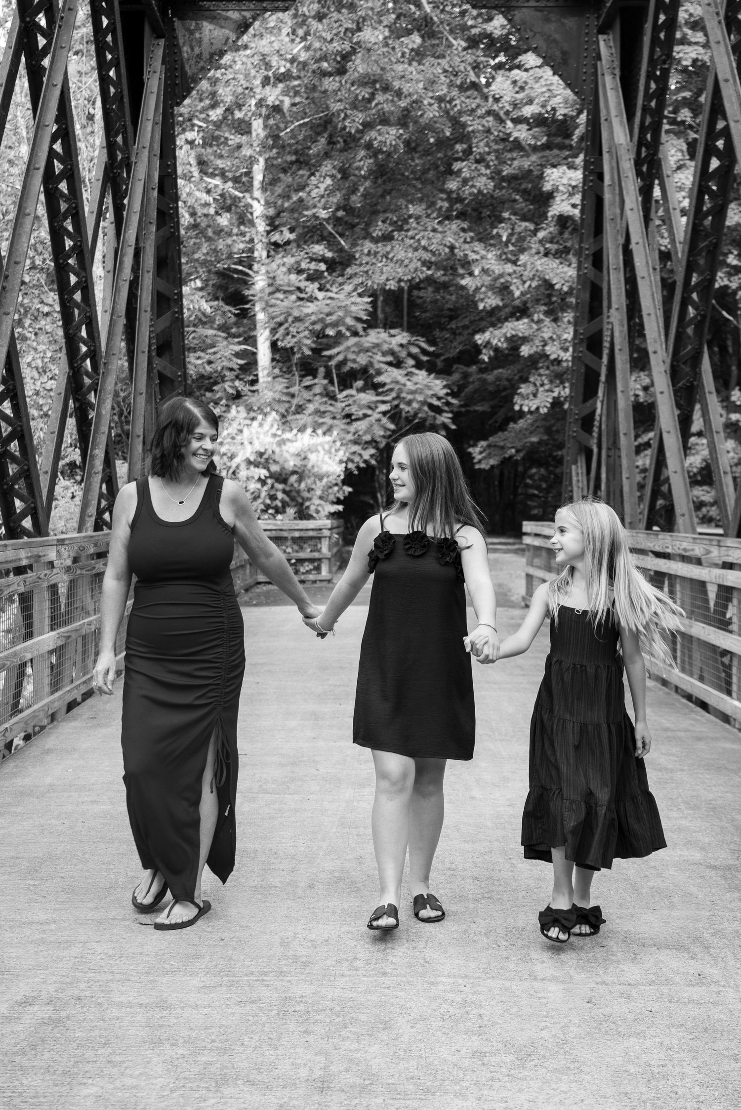Three females in black outfits hold hands walking on a bridge surrounded by lush trees in a black and white photo.