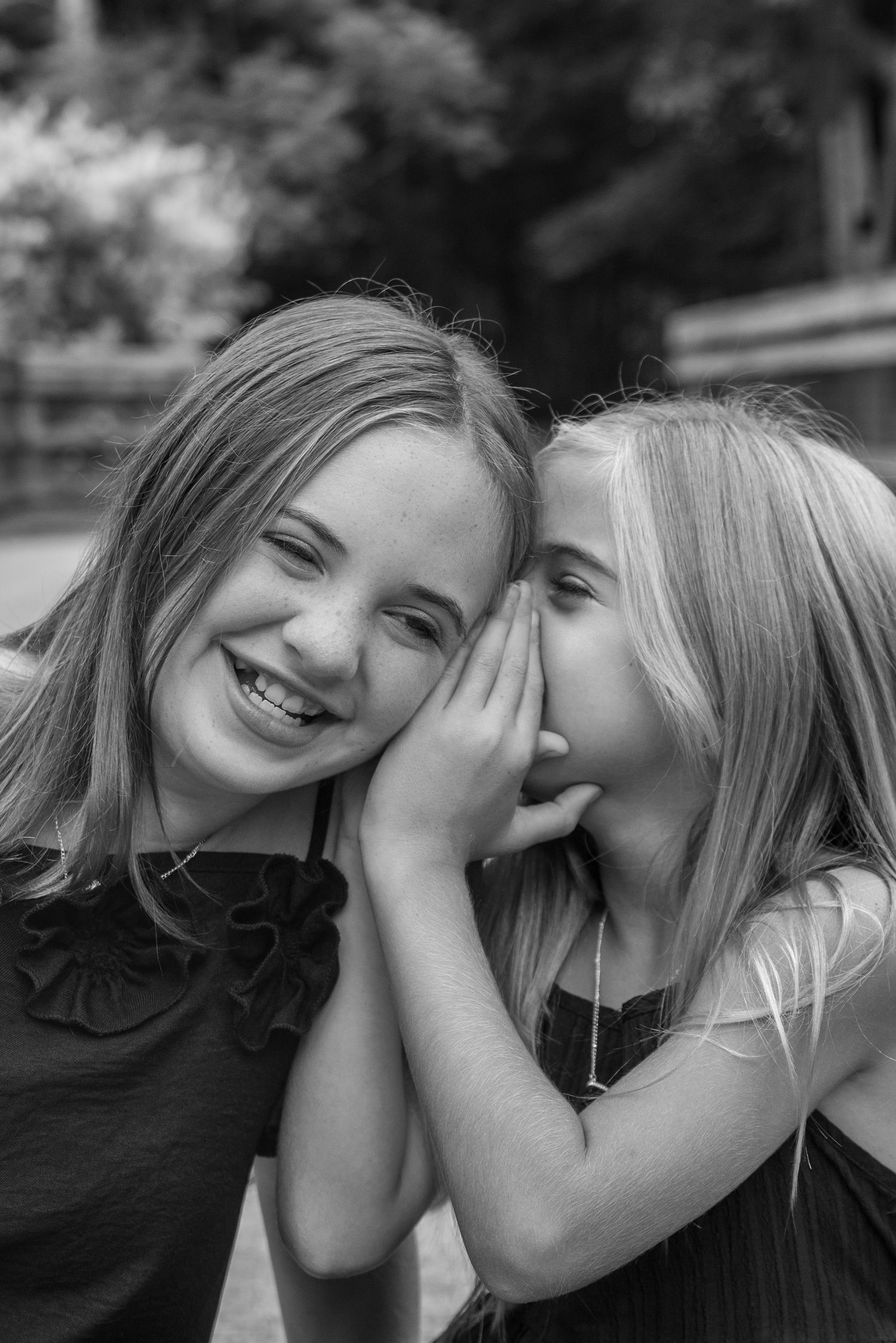 Two blonde girls sharing a secret outdoors, one whispering in the other's ear while she laughs, black and white photo.