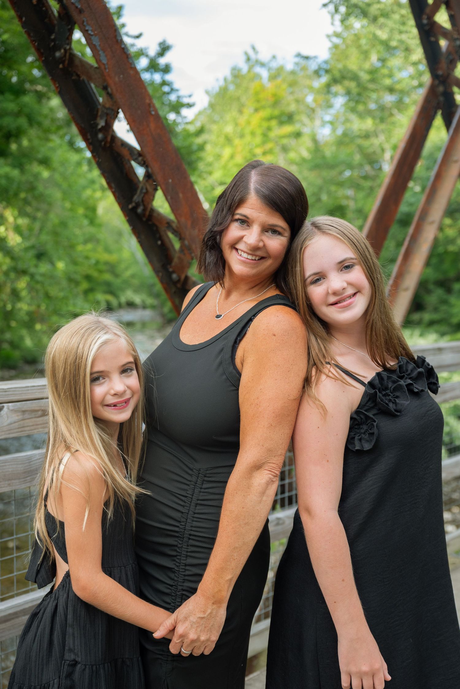 A smiling brunette woman poses with two blonde girls in black dresses on a rustic bridge surrounded by green trees.