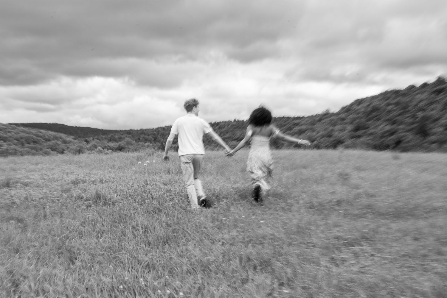 Black and white photo of two children holding hands running through a grassy meadow under cloudy skies.