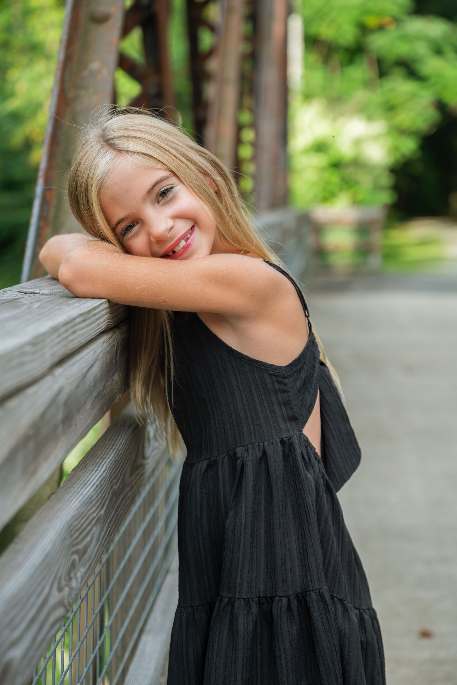 Smiling blonde girl in black dress leaning on wooden bridge railing in a lush green park setting.