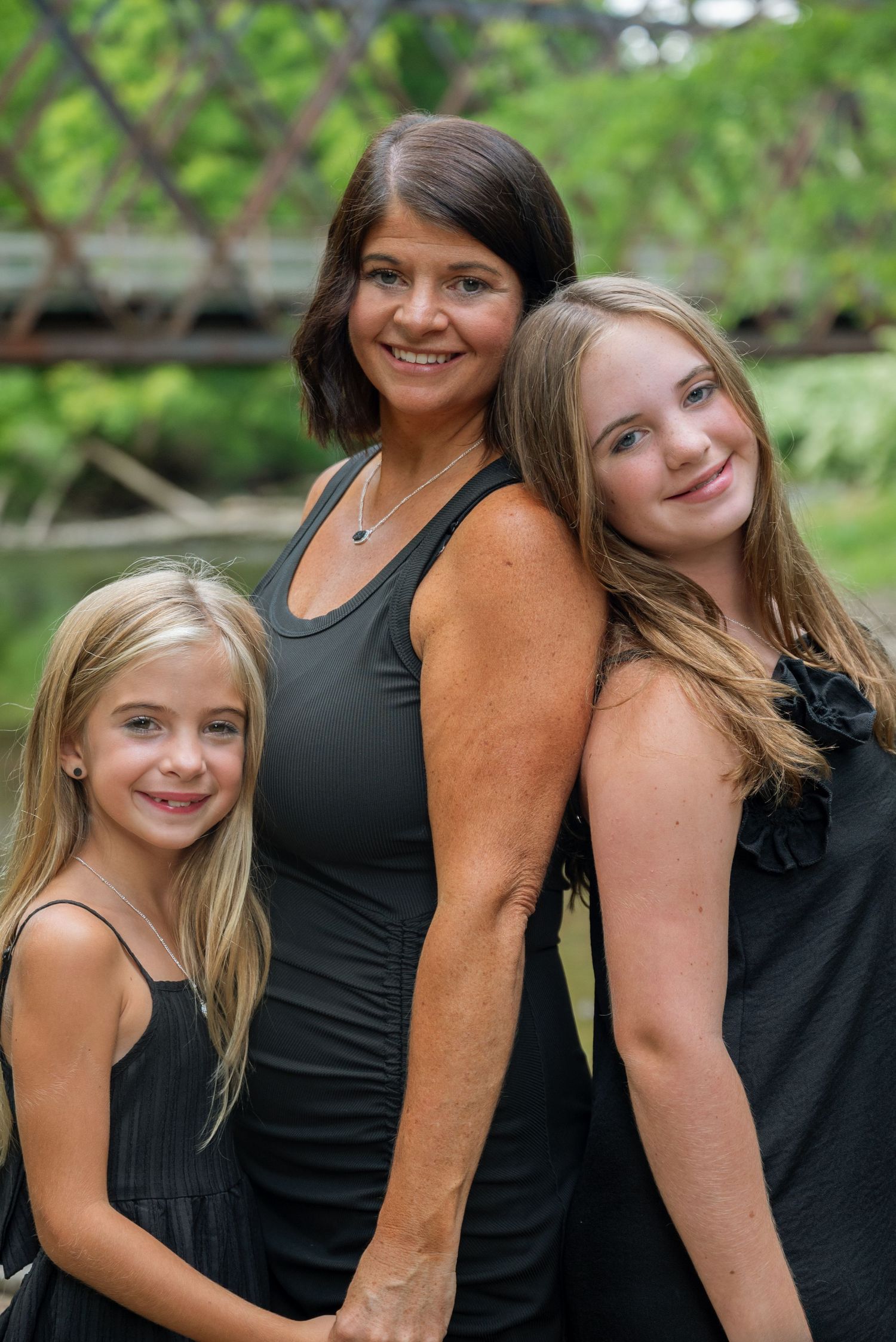 A woman and two blonde girls in matching black outfits smile together outdoors near a bridge.