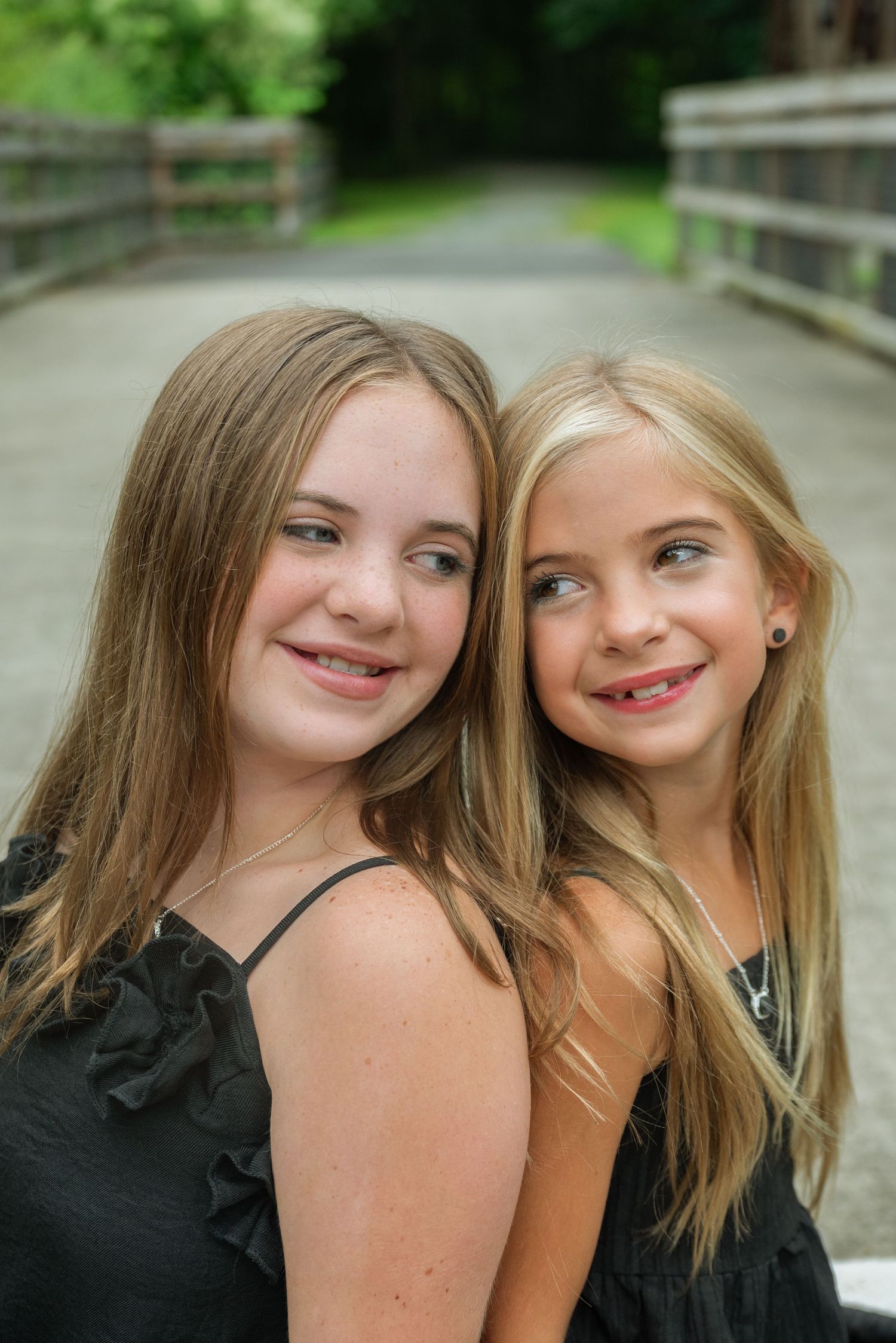 Two smiling girls with long hair wearing black dresses pose together on a wooden bridge outdoors.