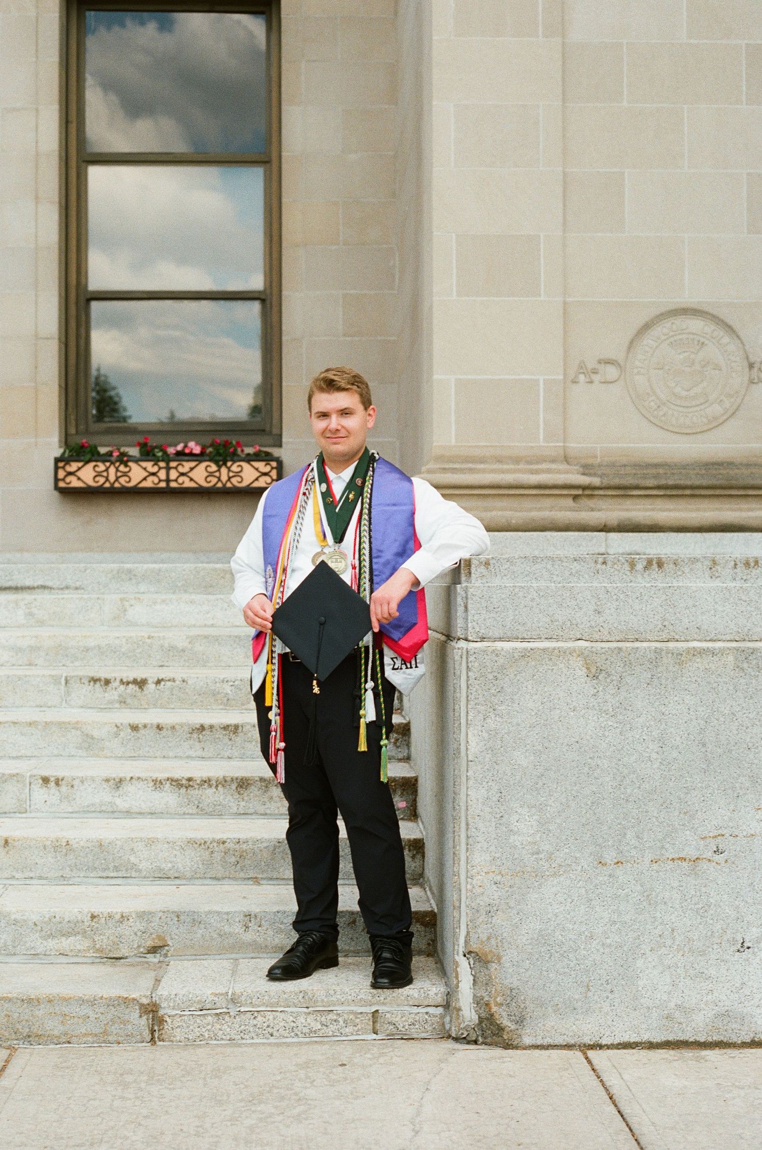 Young man in graduation regalia holding cap on stone steps of academic building.