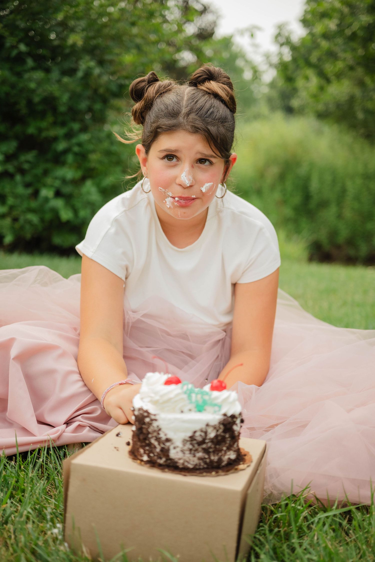 Young girl in pink tulle skirt with frosting on face poses with birthday cake outdoors on grass.