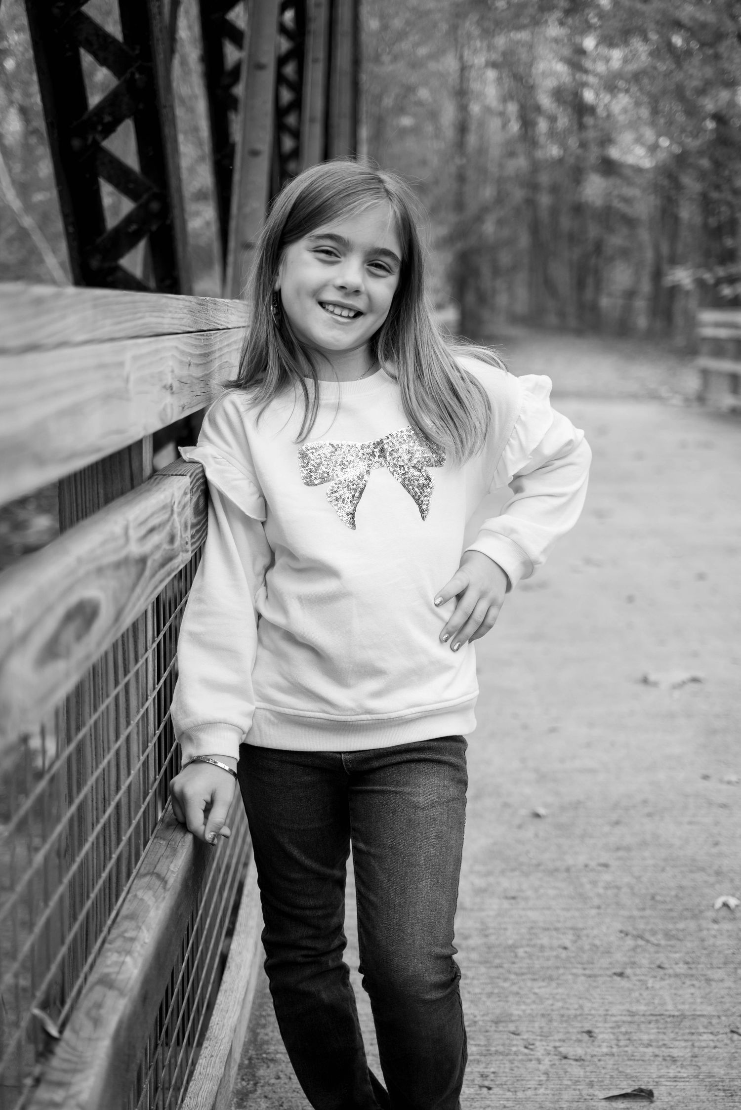 Black and white portrait of a smiling girl in bow sweatshirt leaning on a bridge railing.