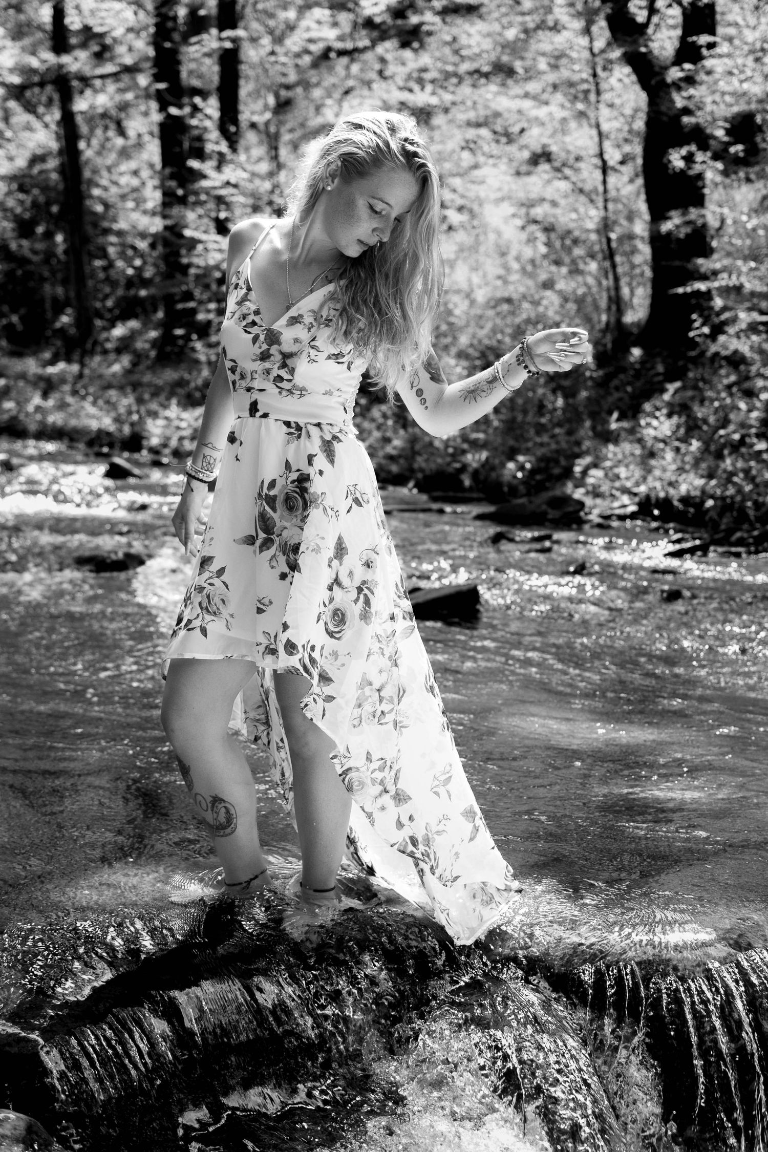 Blonde woman in floral dress wading in a forest stream, black and white photography.