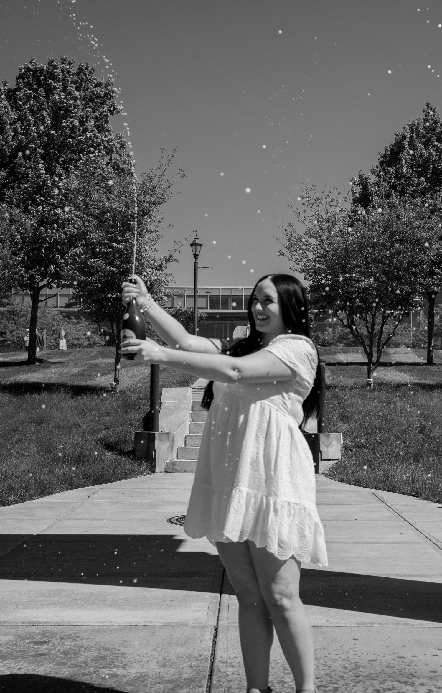 A smiling woman in a white dress pops champagne outdoors in a black and white photo with trees in the background.