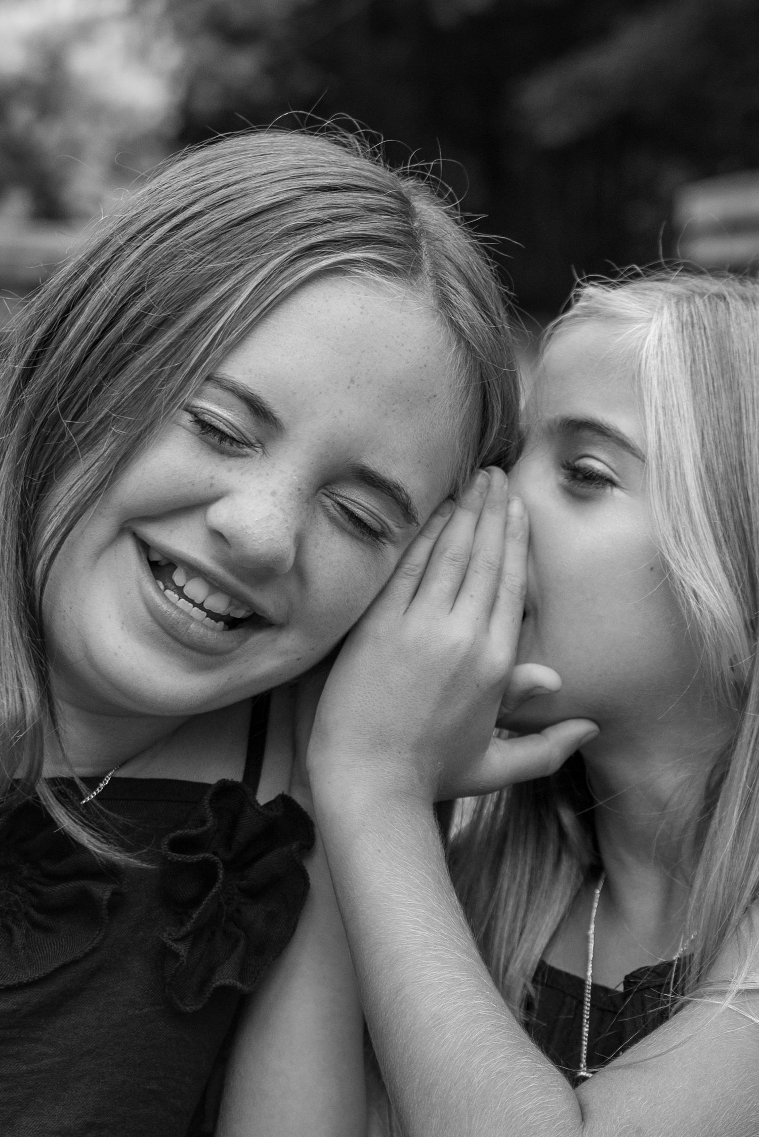 Two young blonde girls sharing a secret whisper, one smiling joyfully, captured in black and white photography.