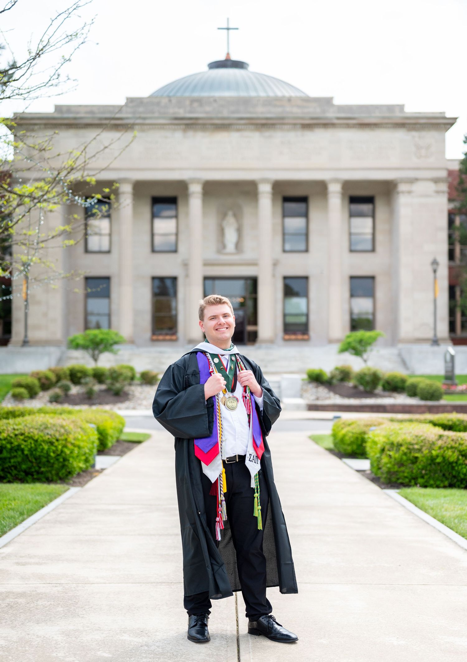 Graduate in black academic regalia with honor cords poses in front of a neoclassical university building.