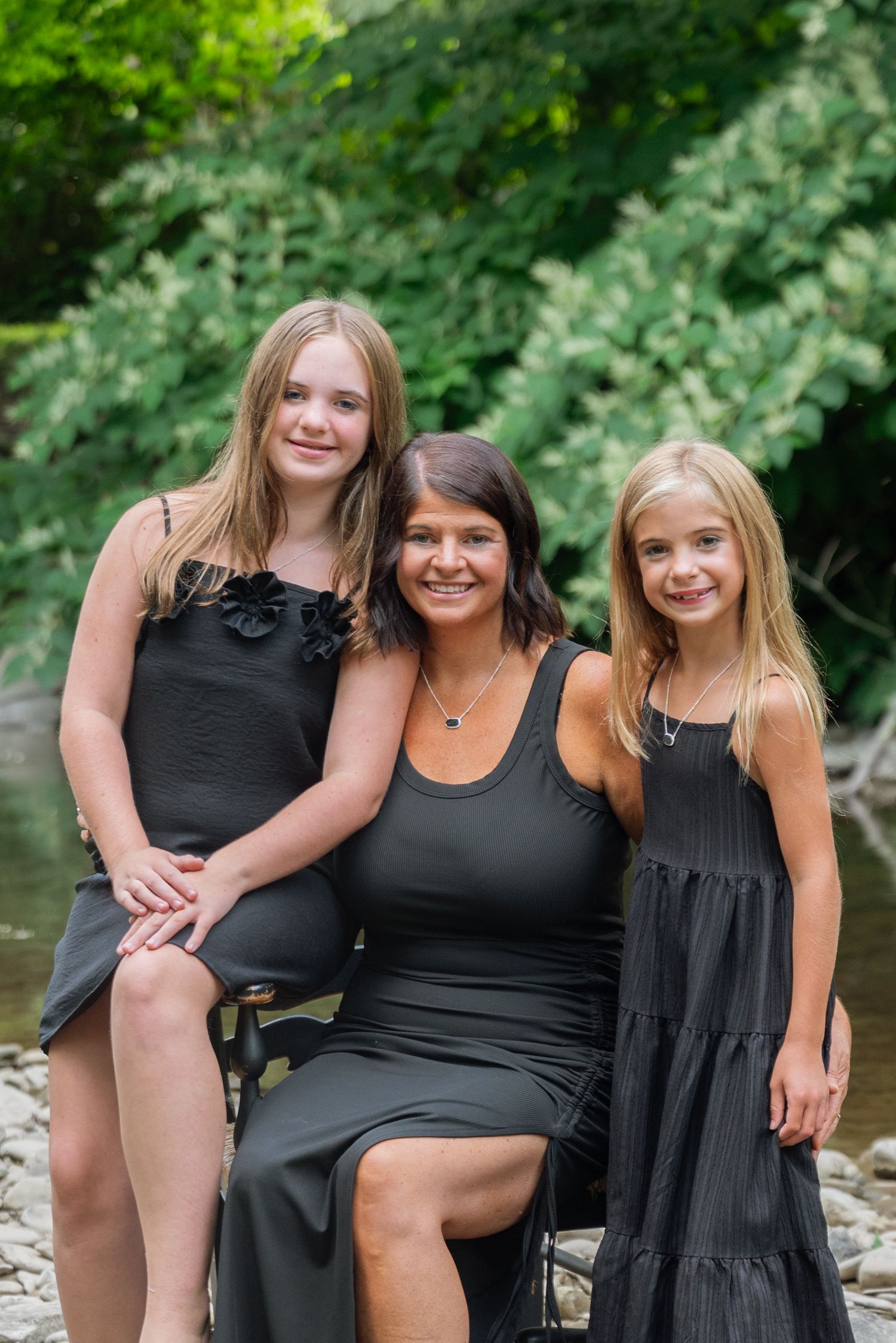 A woman and two blonde girls in matching black outfits pose together outdoors near a creek surrounded by green trees.