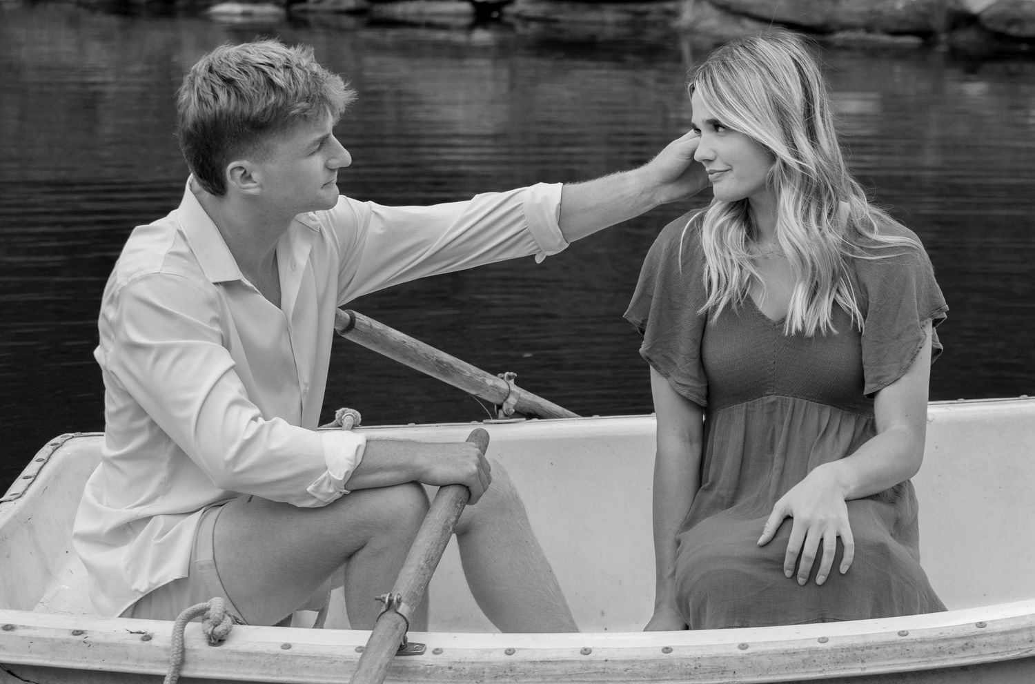A couple shares a romantic moment in a rowboat, with the man gently touching the womans hair near a serene lake.