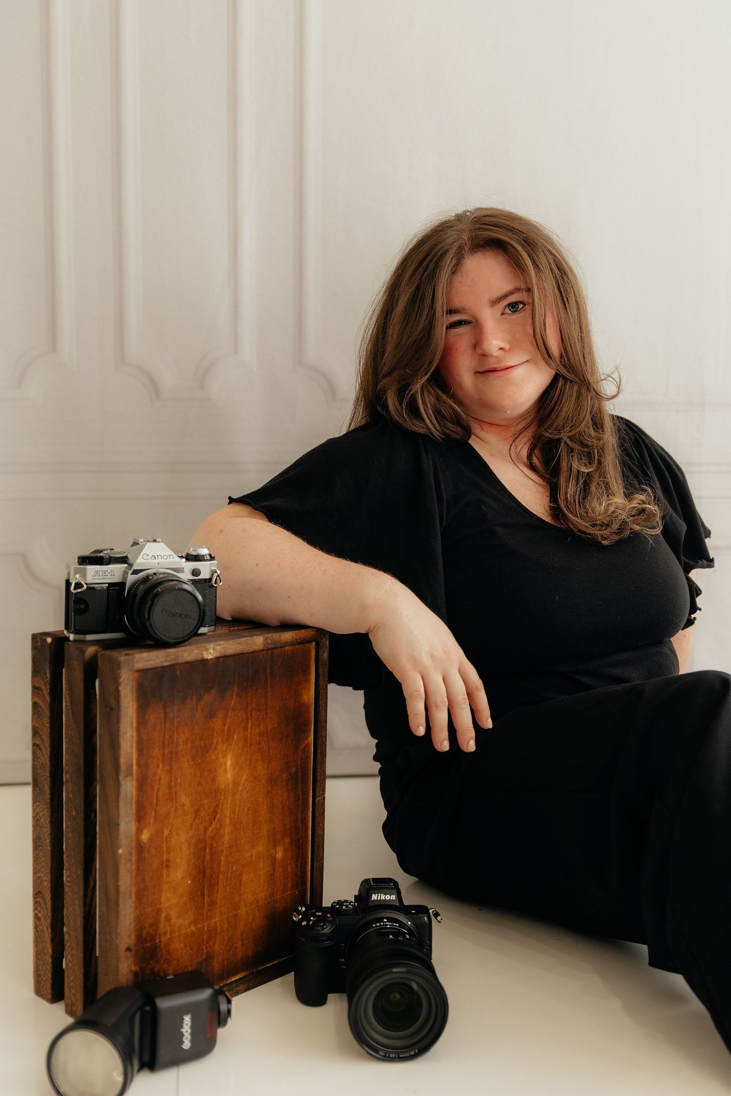 Woman in black dress poses with vintage and modern cameras on wooden crates in a white studio setting.