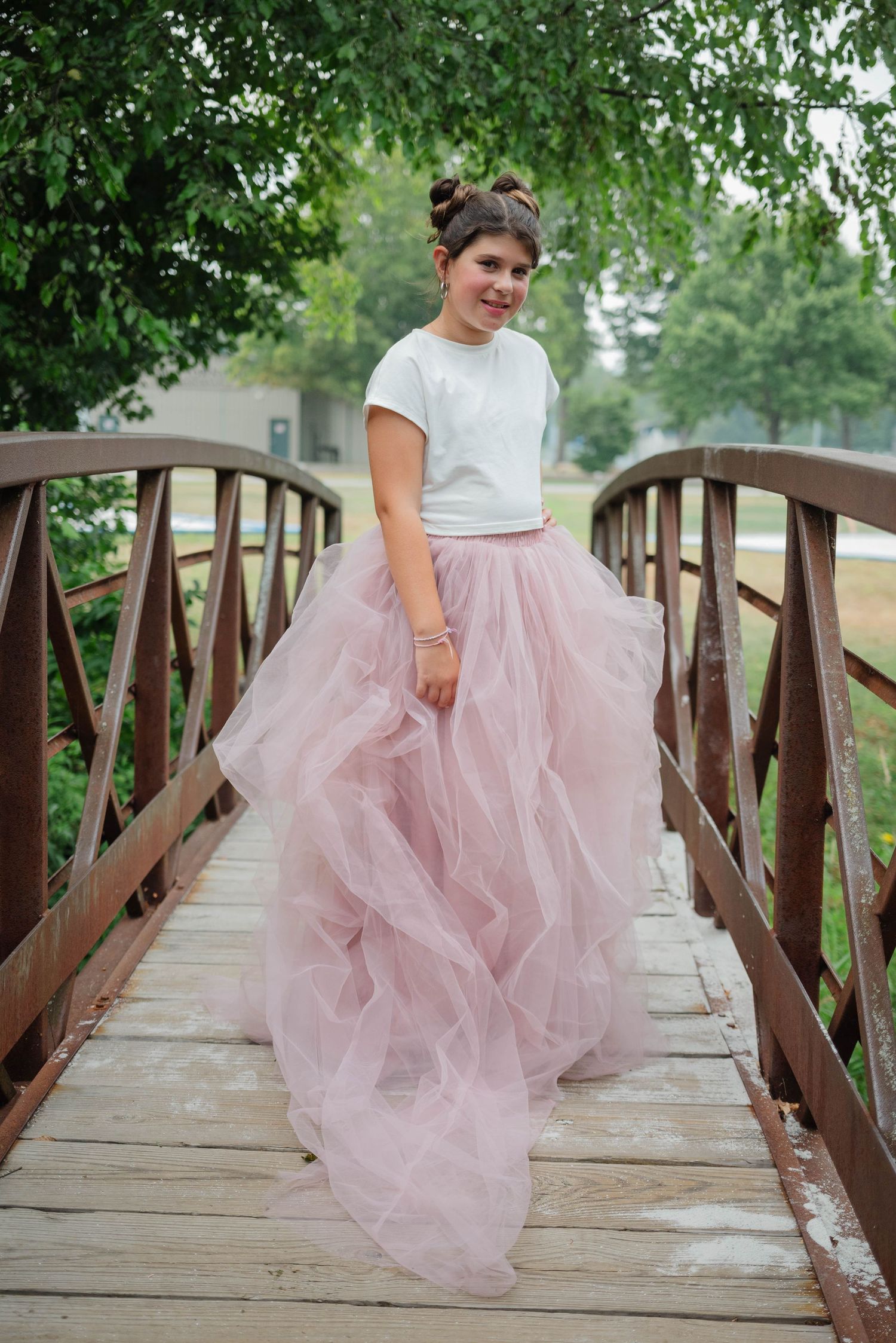 Young girl in white top and pink tulle skirt standing on a wooden bridge surrounded by greenery.