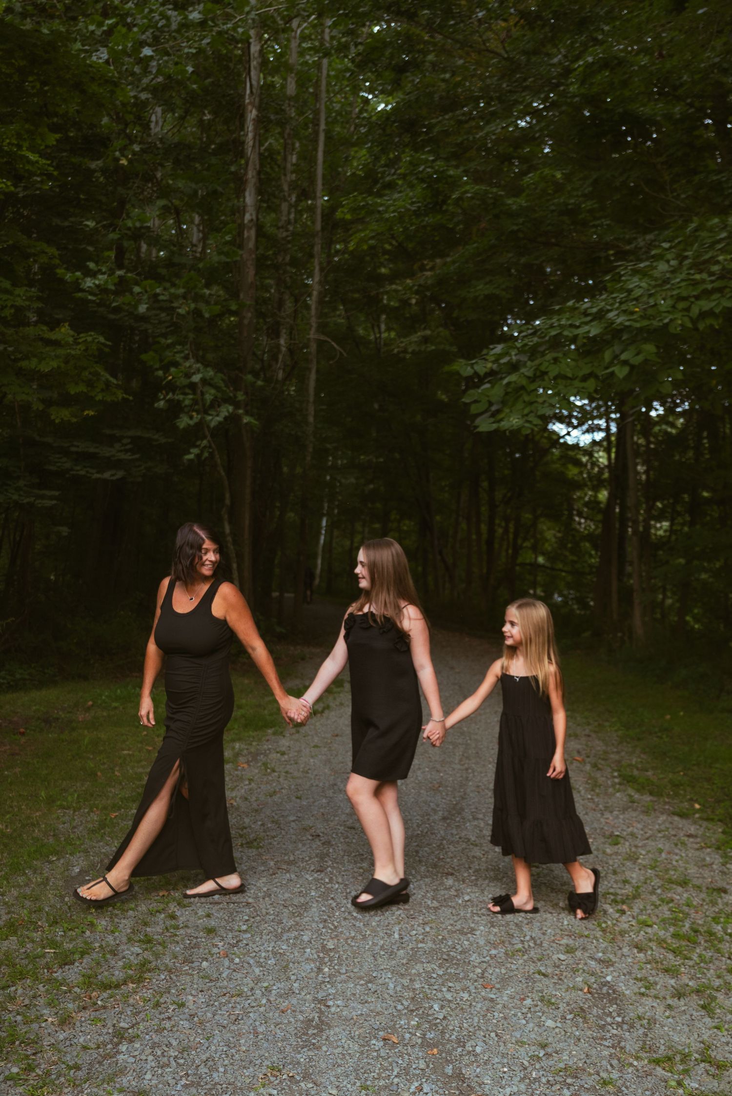 Three females in matching black dresses hold hands on a forest path, posing for a family portrait.