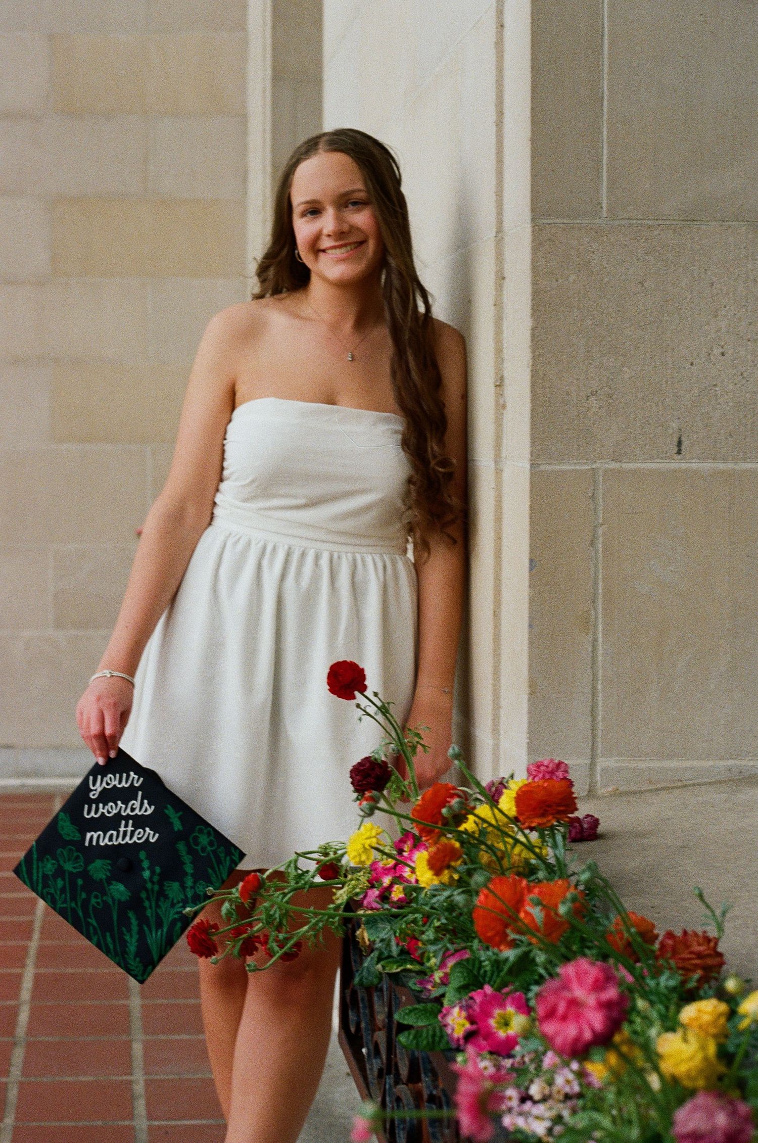 Smiling graduate in white dress holds decorated cap reading 'your words matter' beside colorful flowers.