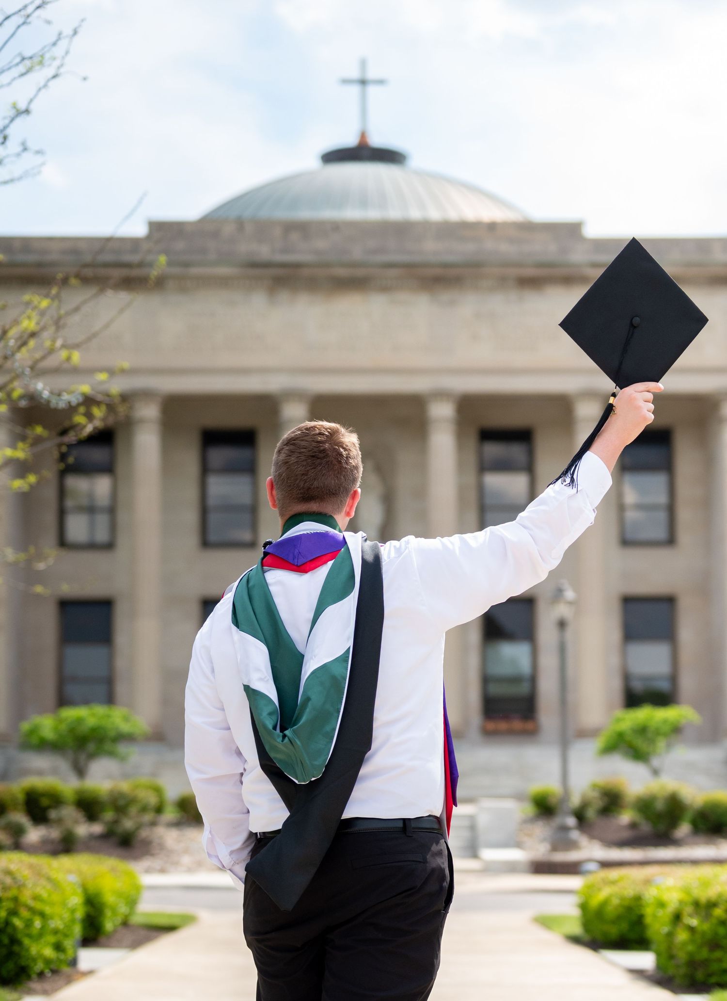 Graduate raises cap toward domed university building with cross, wearing academic regalia, celebrating commencement outdoors.