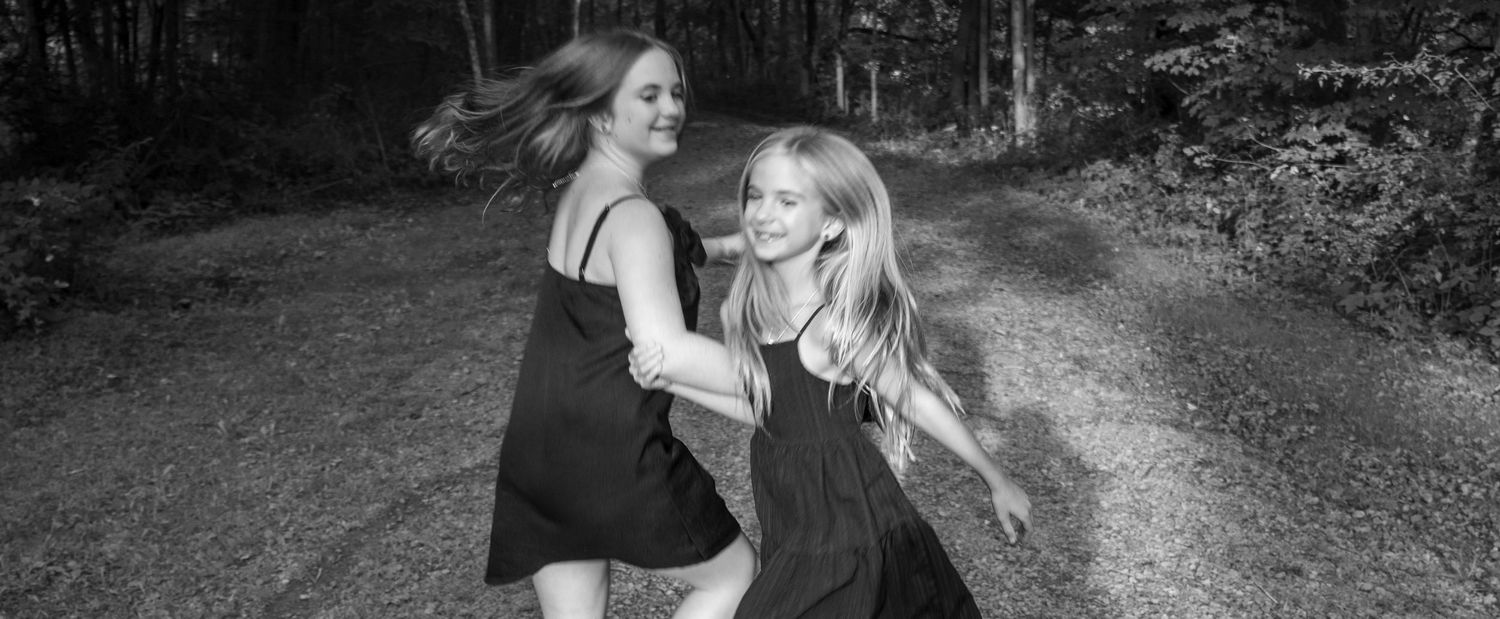 Two young girls in black dresses laugh and run together on a forest path in a black and white photo.