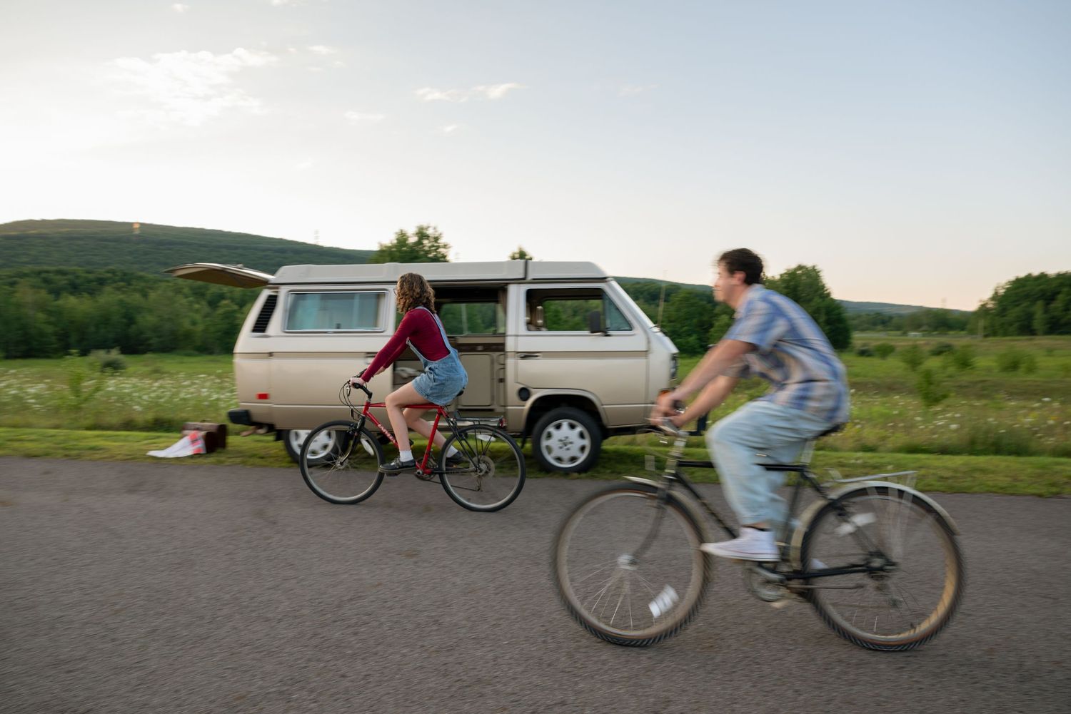 Two cyclists ride bikes past a vintage VW camper van parked on a scenic road surrounded by green fields.