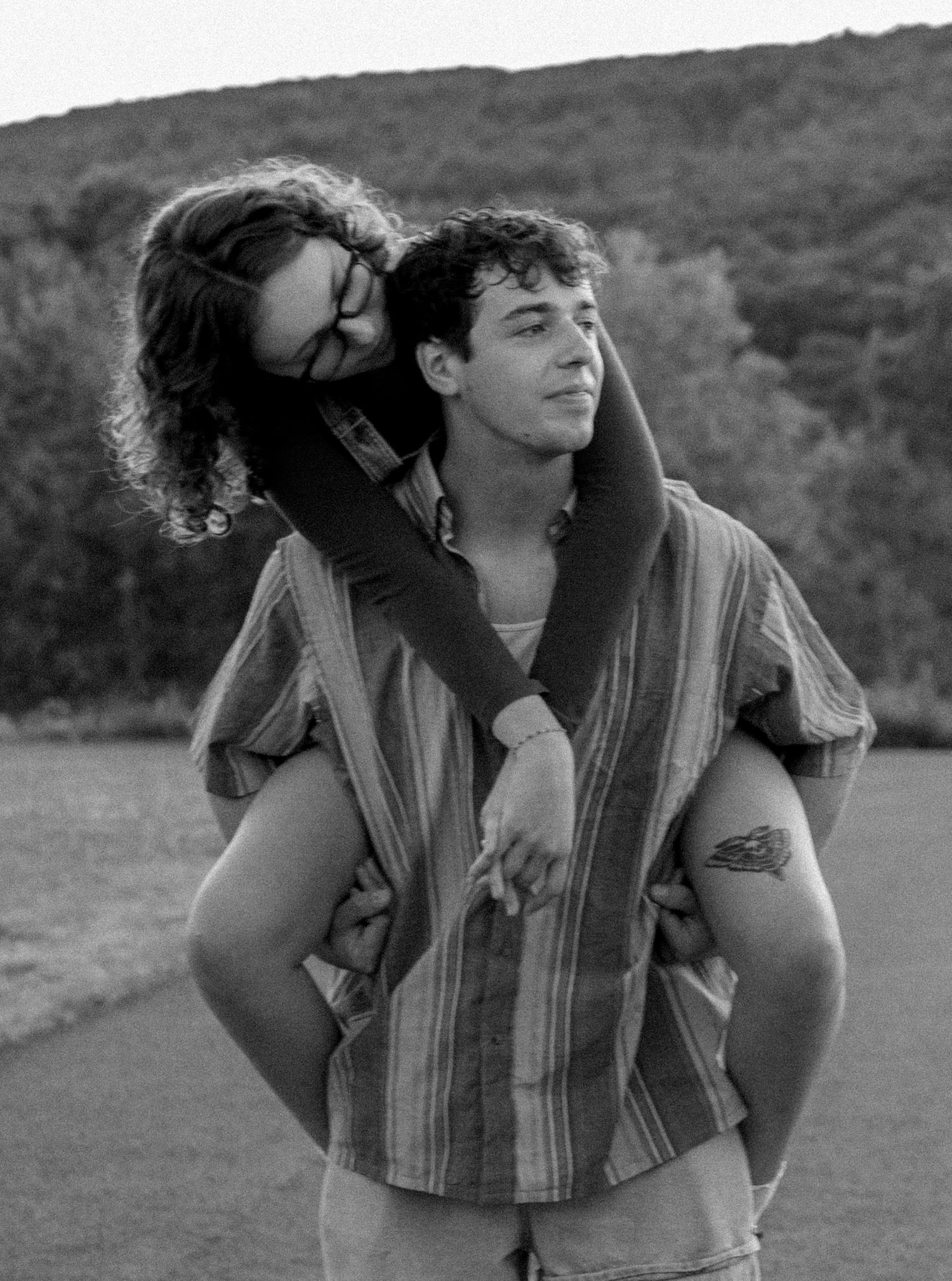 Black and white photo of a young couple, woman giving a piggyback ride to a man outdoors with hills in background.