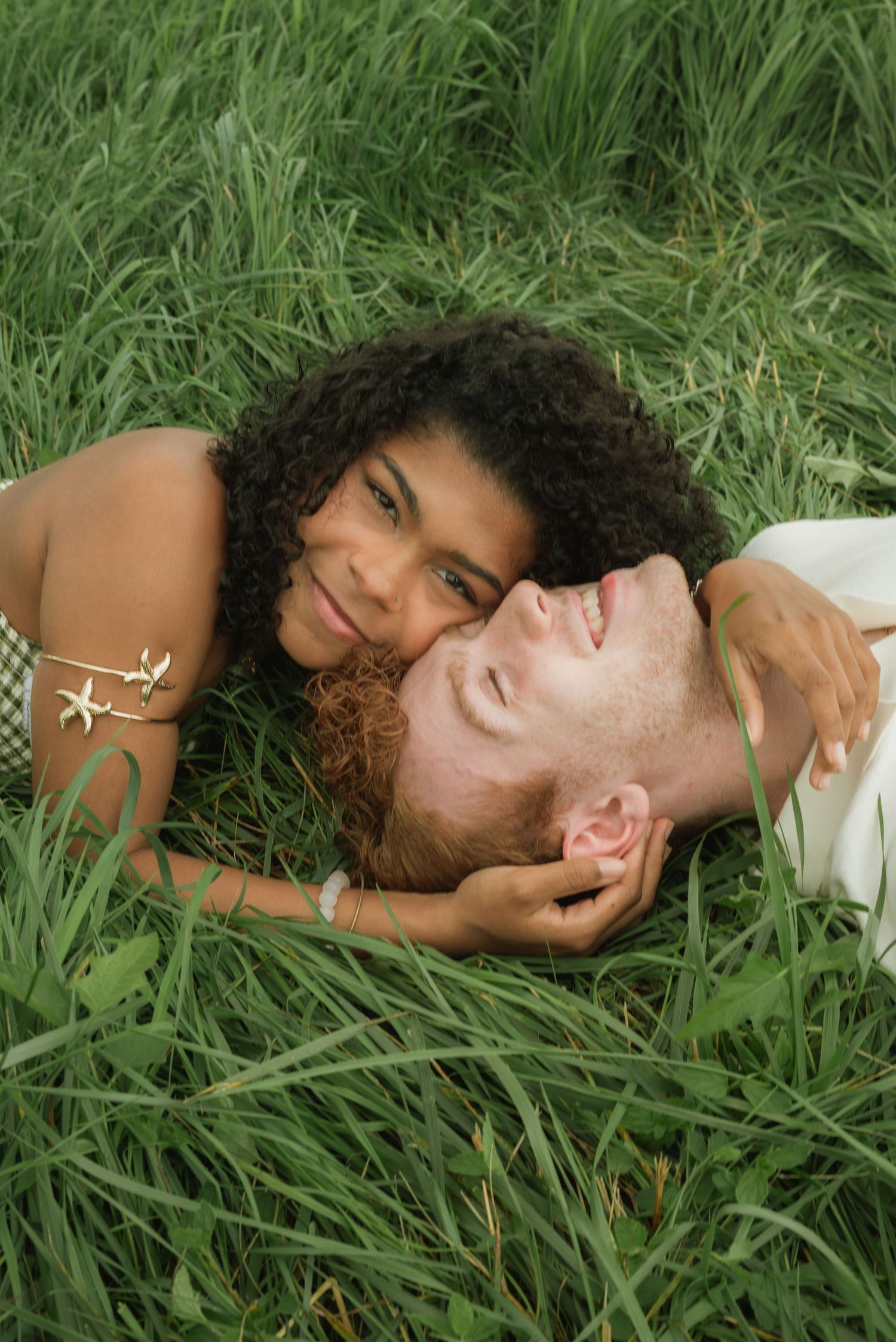 A couple lying in lush green grass, smiling and embracing in a romantic outdoor moment.