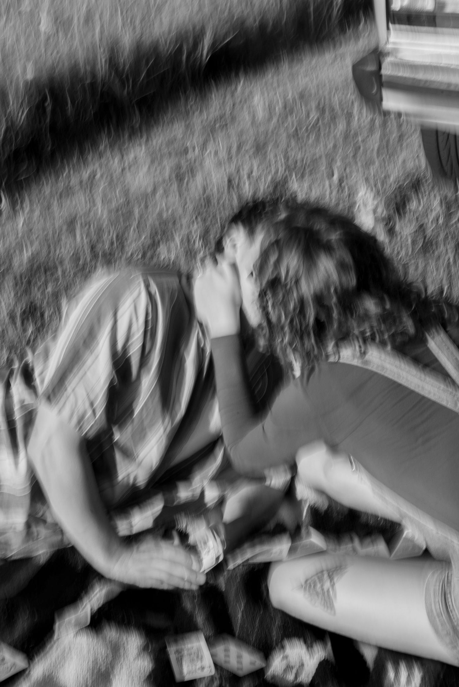 Black and white photo of two people sitting outdoors on grass, leaning close together near a vehicle.
