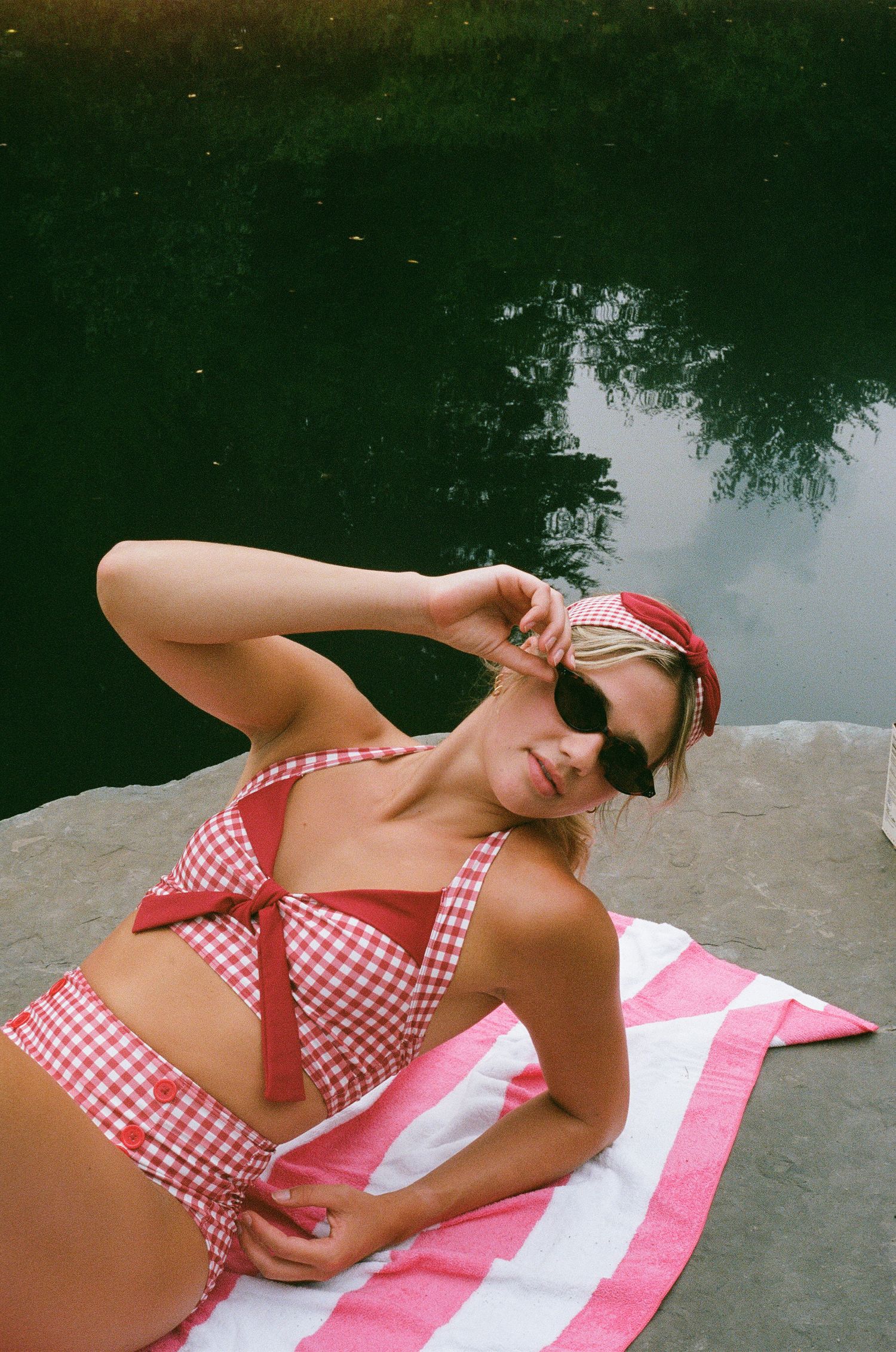 Woman in red gingham bikini and sunglasses lounging on a striped towel by a reflective pond.