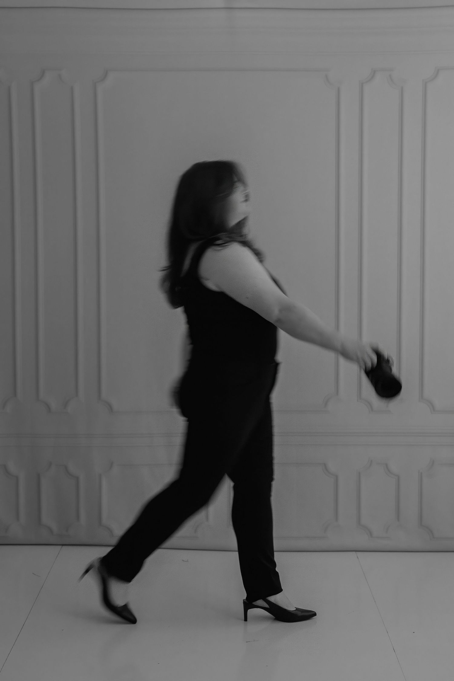 Black and white motion blur photo of a woman in black outfit and heels walking dynamically against ornate white wall.