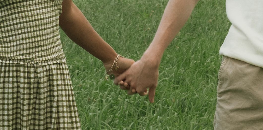 Couple holding hands in a green grassy field, woman wearing checkered skirt with gold bracelet.