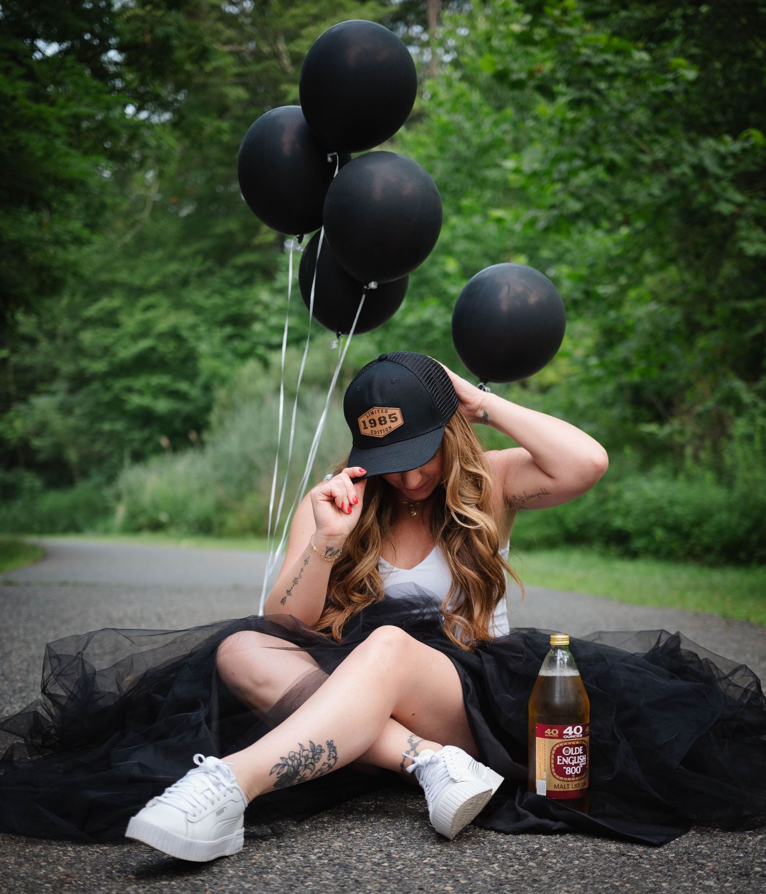 Woman in black tulle skirt sitting on road with black balloons and bottle, cap covering face.