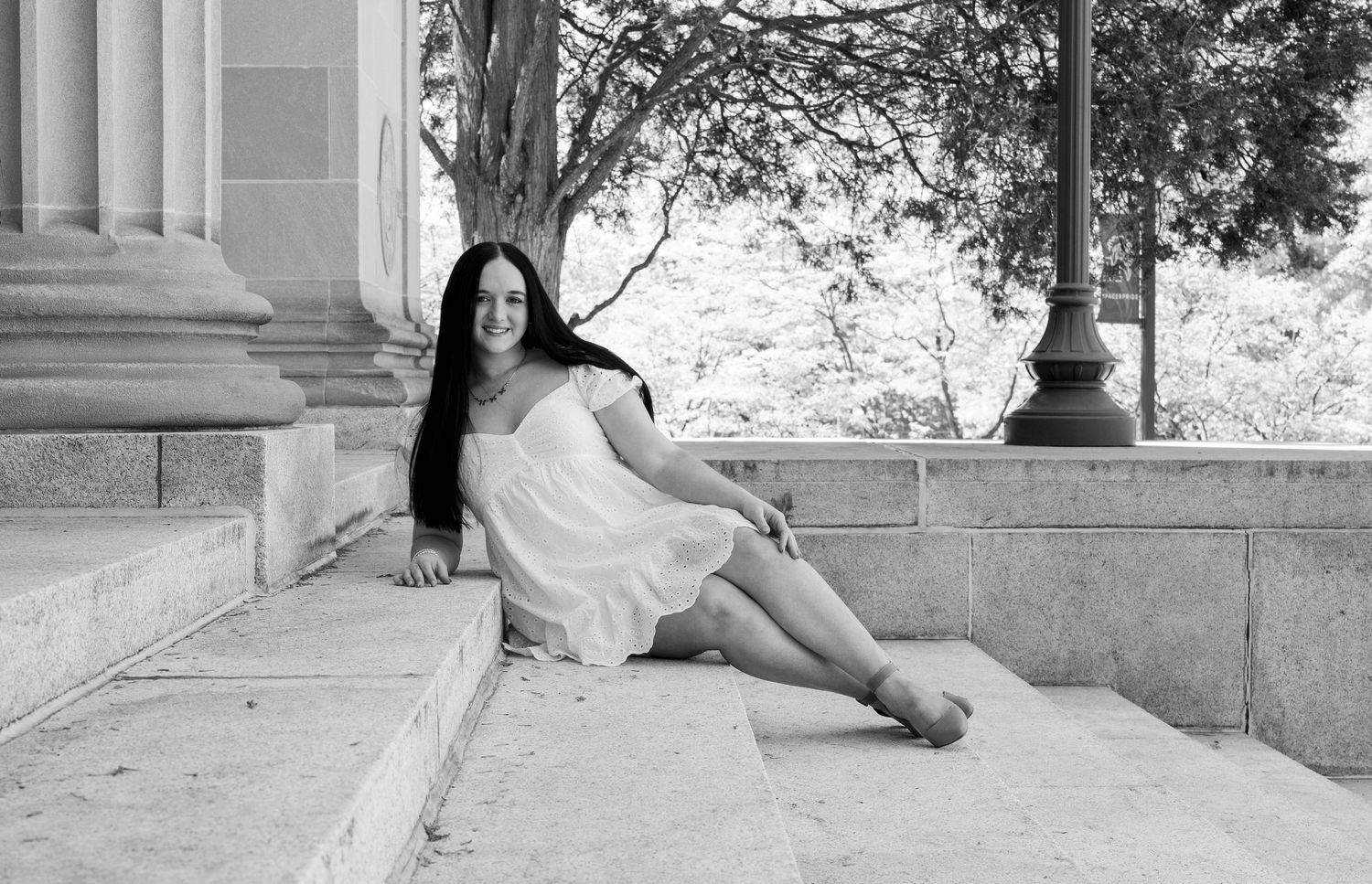 Black and white photo of a smiling young woman in a white dress sitting on stone steps near classical columns.