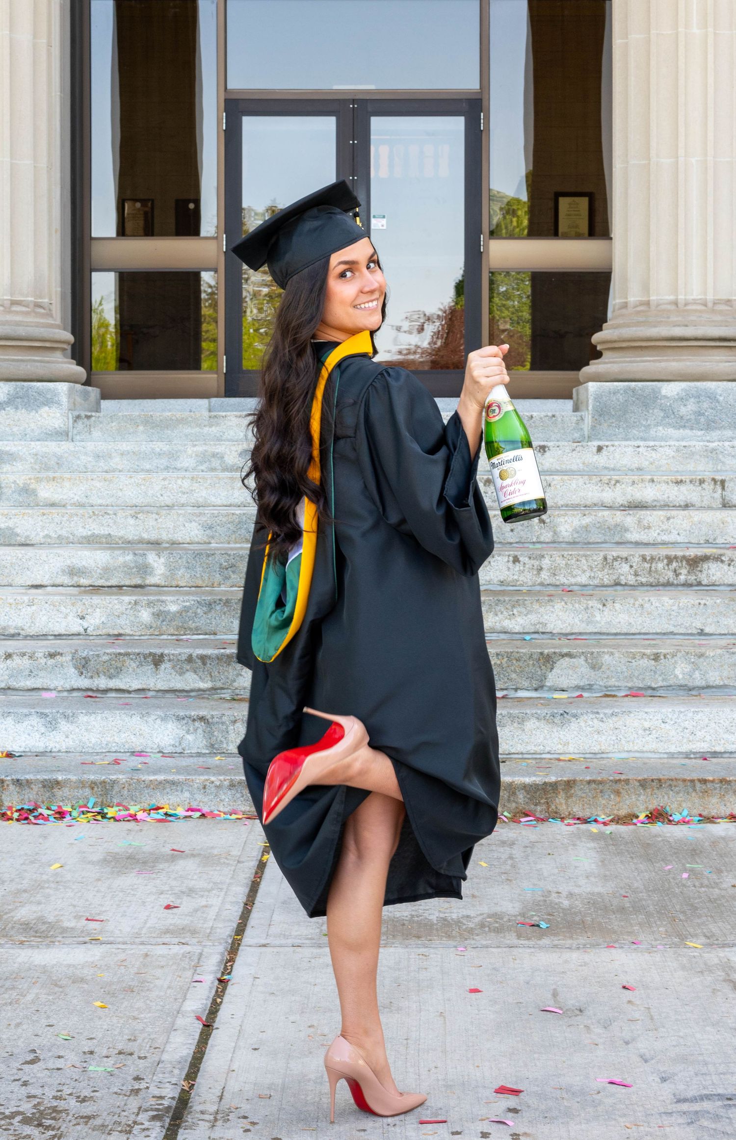 Young woman in black graduation cap and gown holding champagne bottle on university steps, celebrating her graduation.