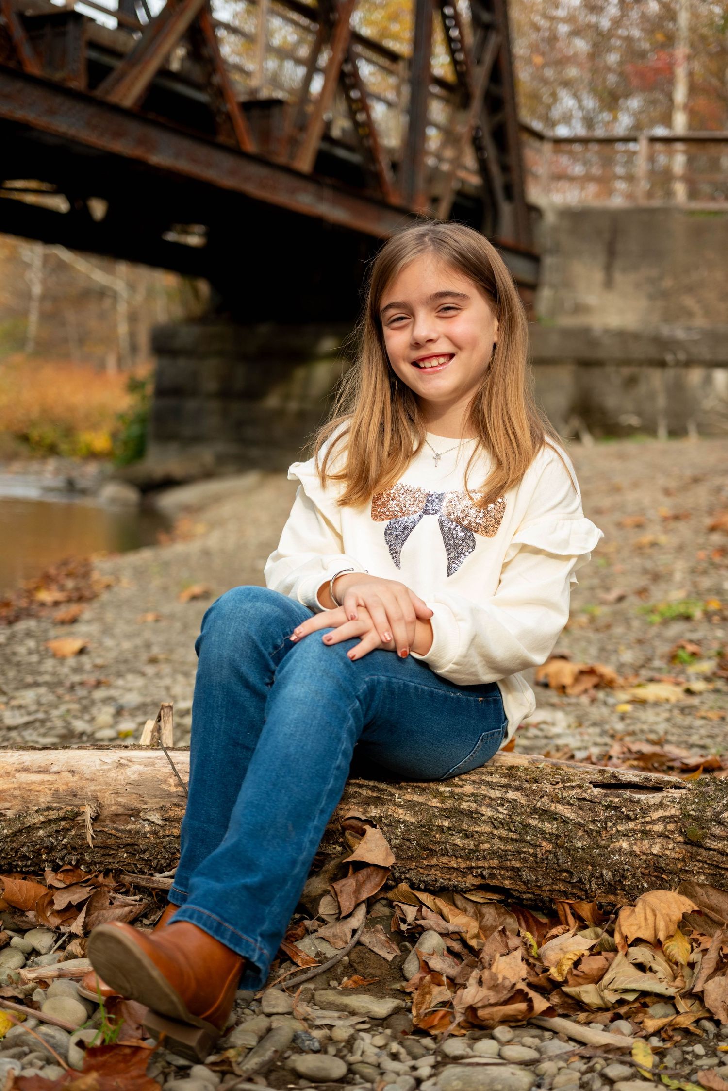 Cheerful young girl in cream sweatshirt and jeans sitting on a log near a rustic bridge in fall.