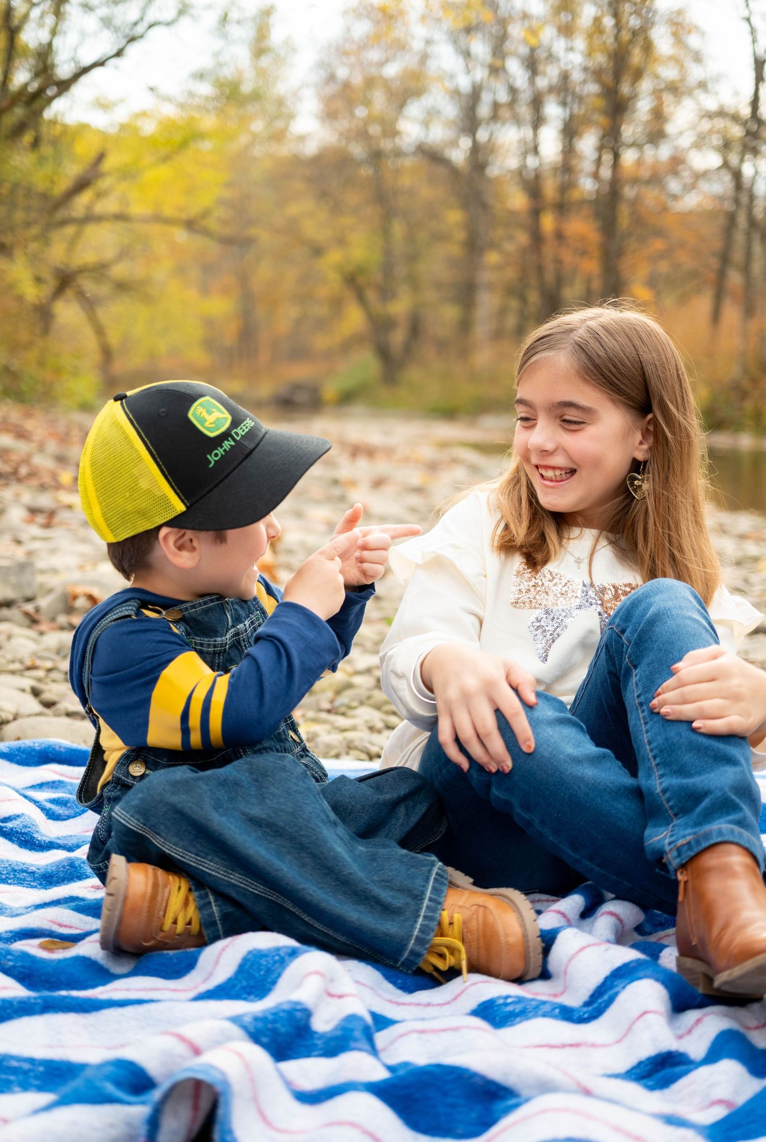 Two children sit on a blanket outdoors in fall, a boy in a John Deere cap pointing at his smiling sister.