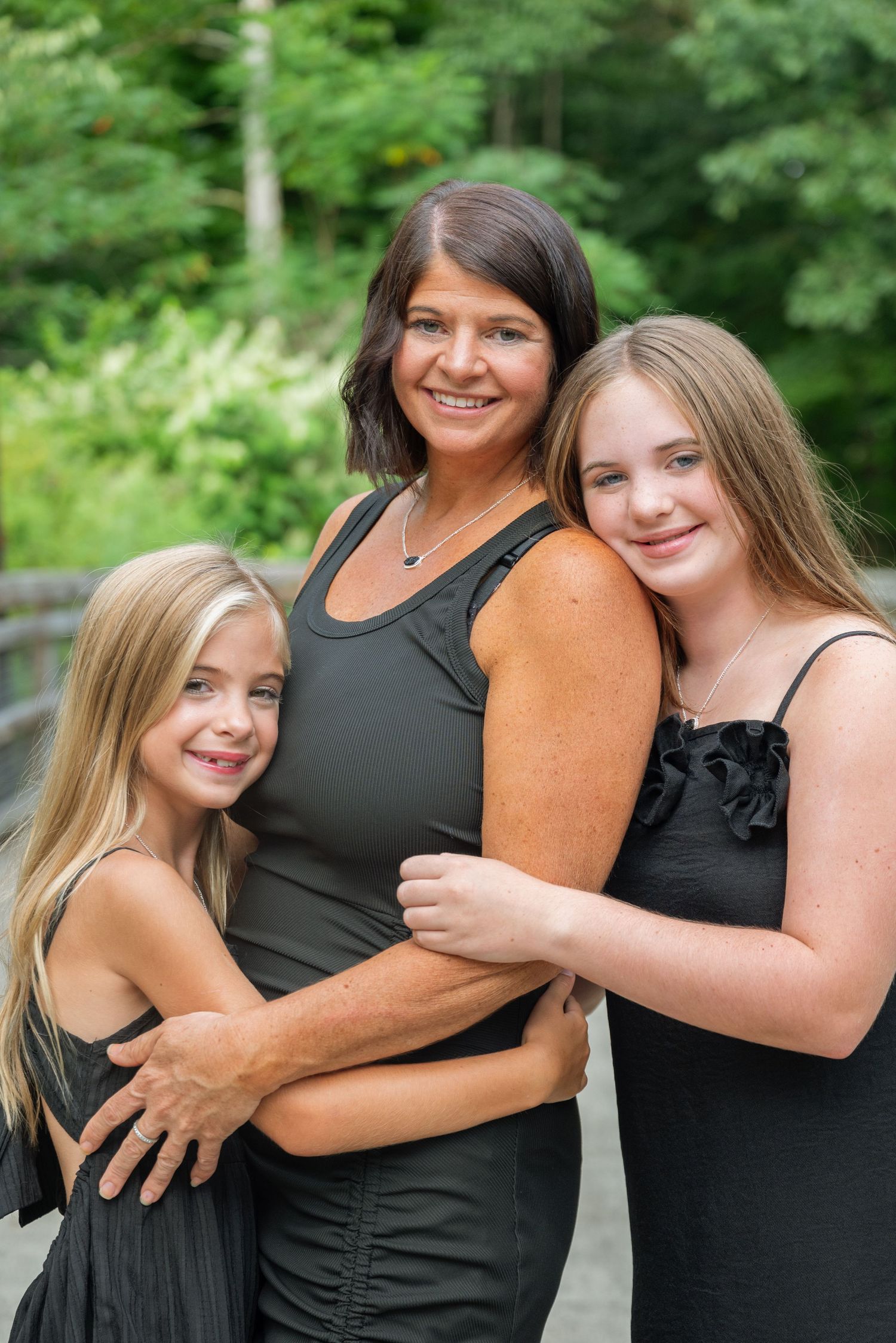 A woman and two girls in matching black outfits smile together outdoors with lush green trees in the background.