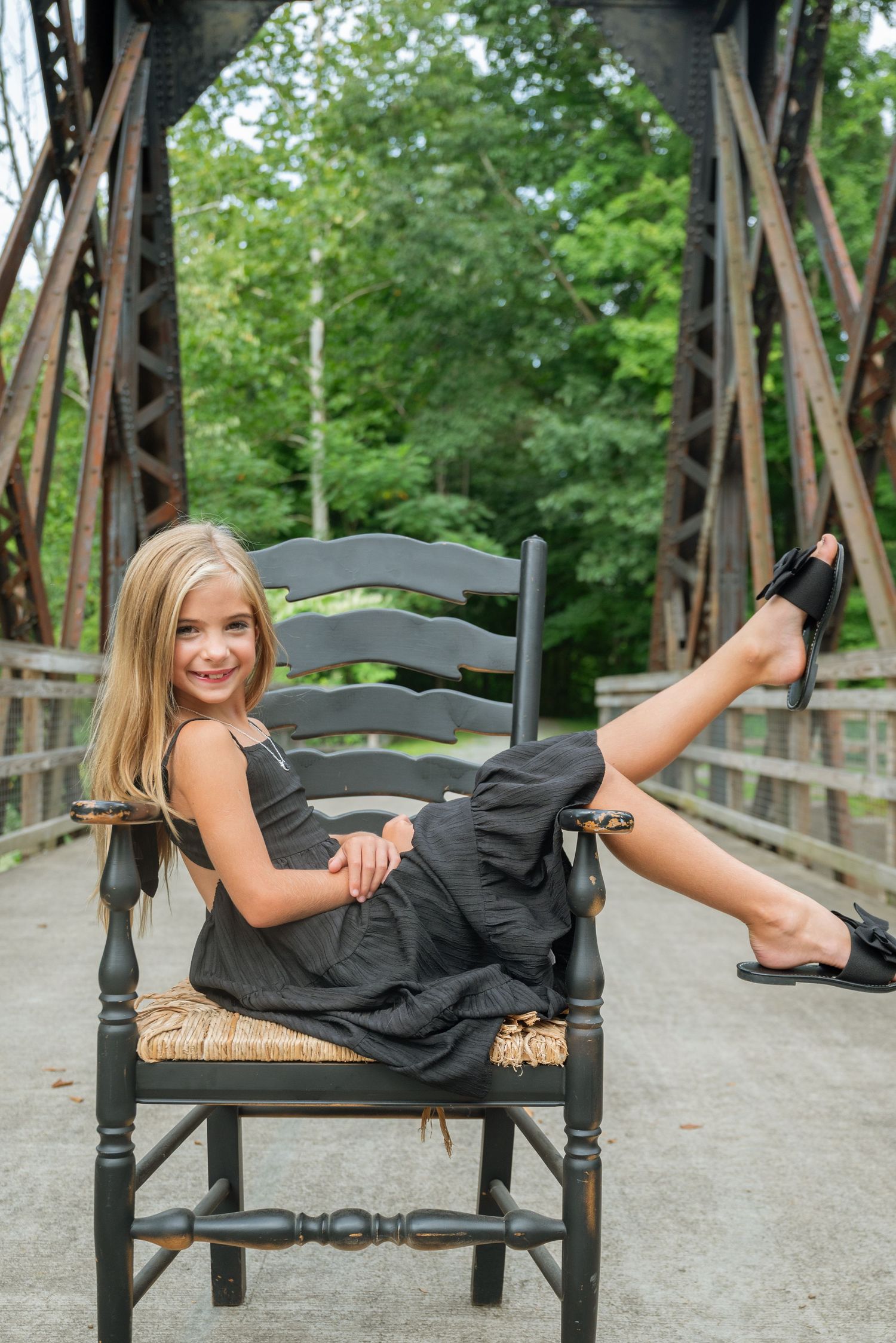 Young girl in black dress sitting relaxed on a dark wooden chair on a rustic bridge surrounded by green trees.
