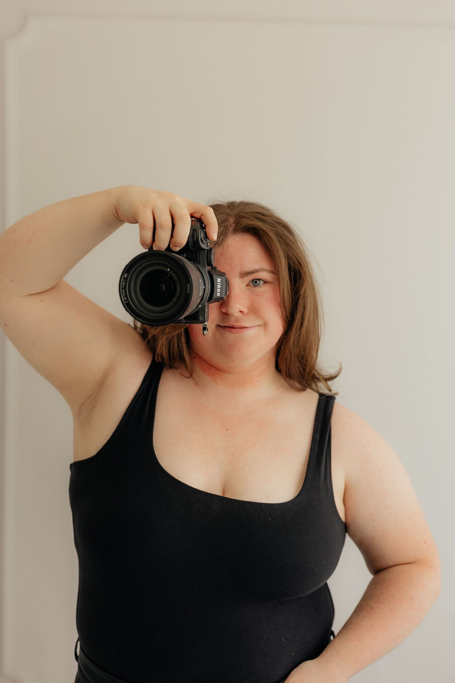 Woman in black bodysuit holding a DSLR camera up to her face, smiling slightly against a white background.