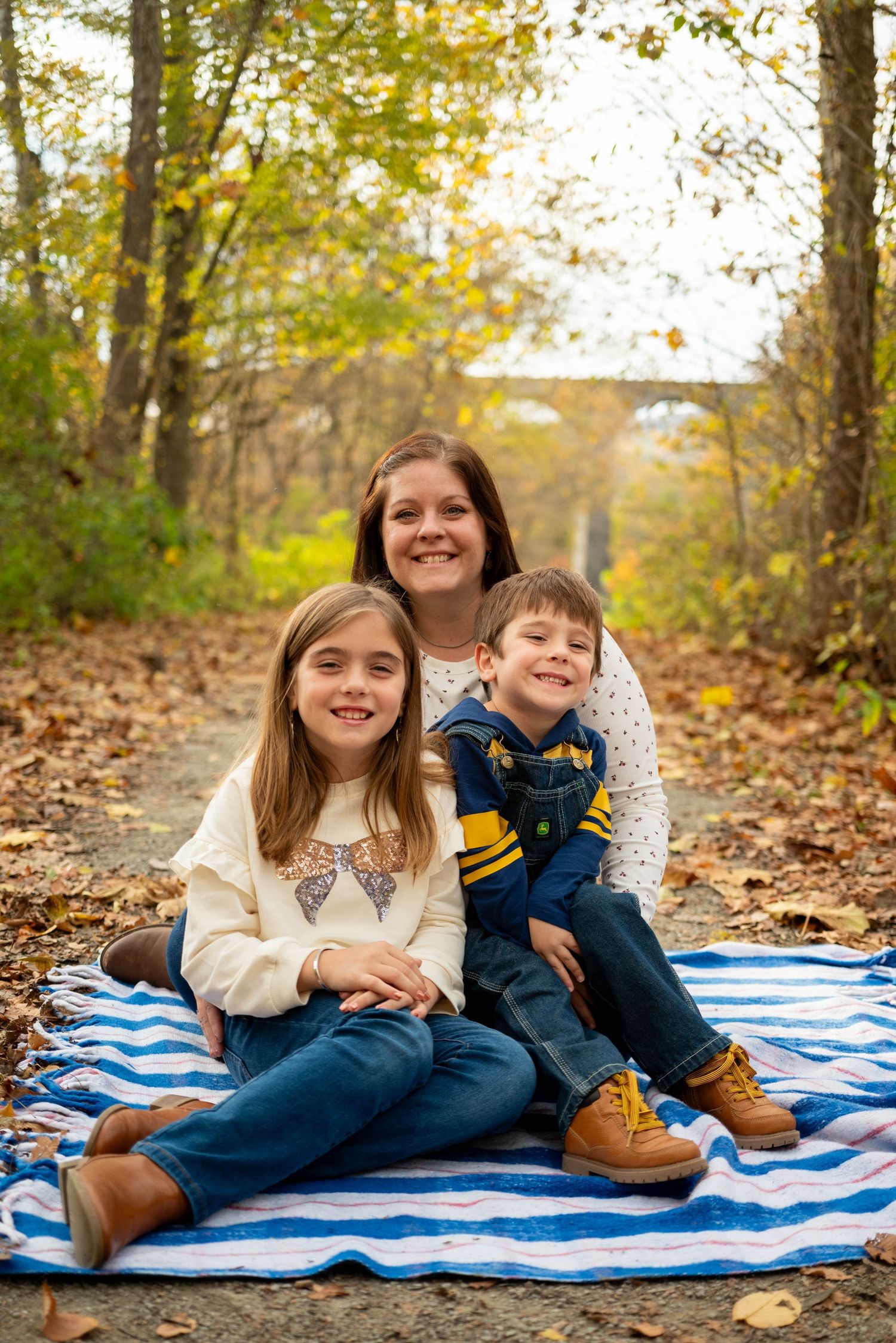 Smiling mother poses with two children on blue blanket surrounded by fall foliage.
