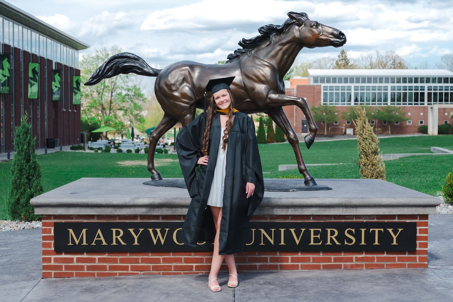 Graduate in cap and gown poses before Marywood University bronze horse statue on campus.