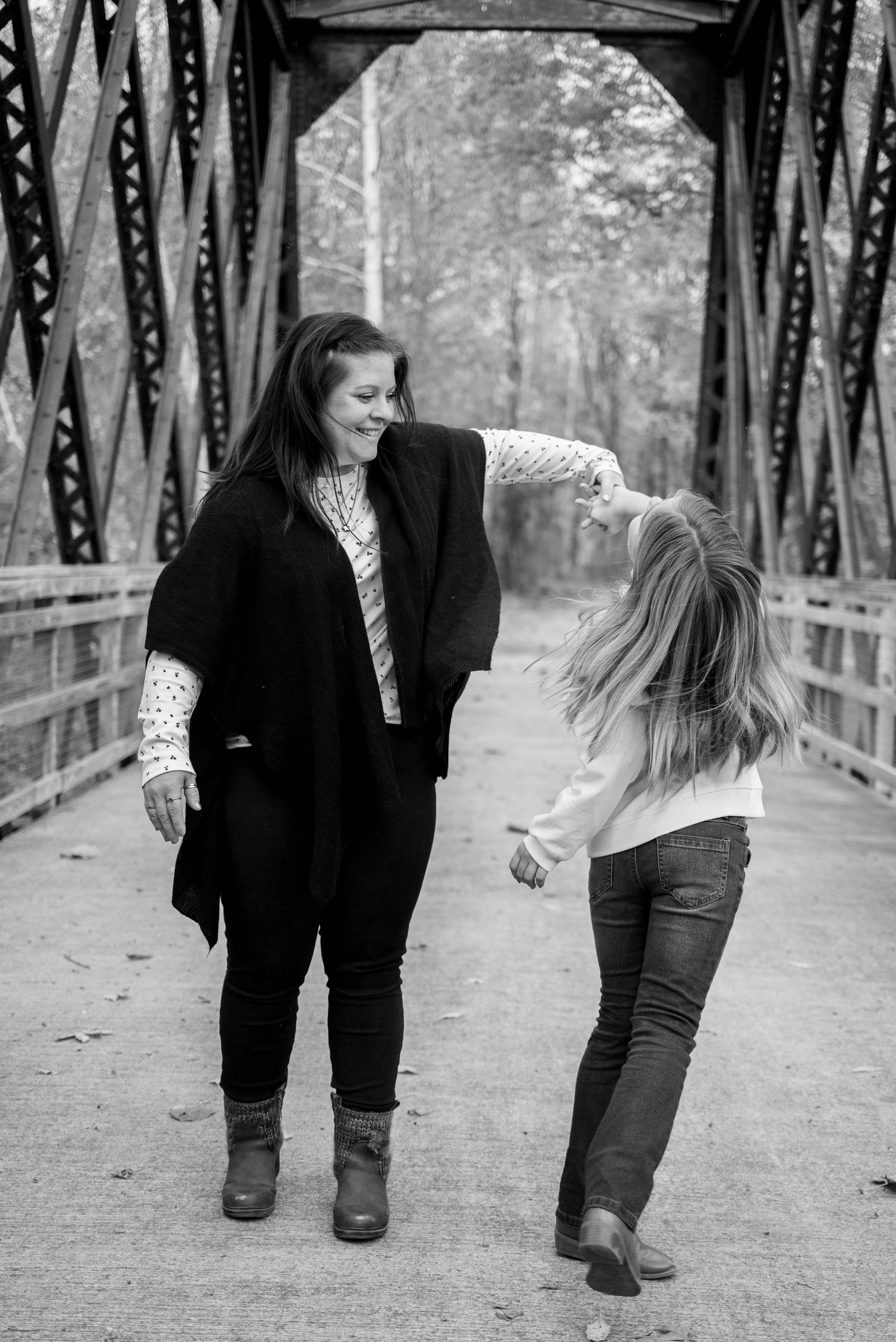 Two women laughing and dancing together on a rustic iron bridge surrounded by trees in a black and white photo.