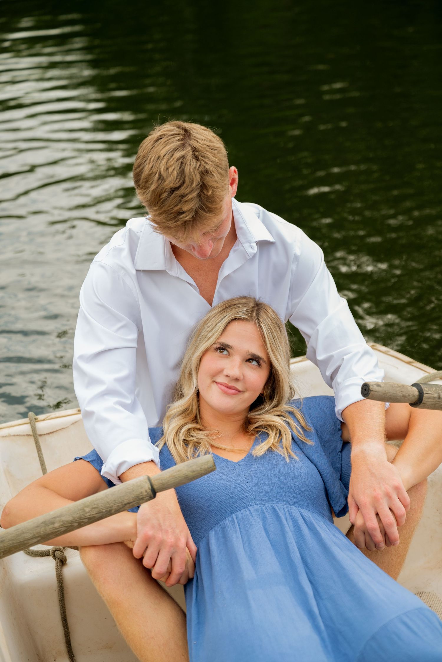 Couple relaxing in a rowboat on a lake, woman in blue dress leaning back while man in white shirt looks down at her.