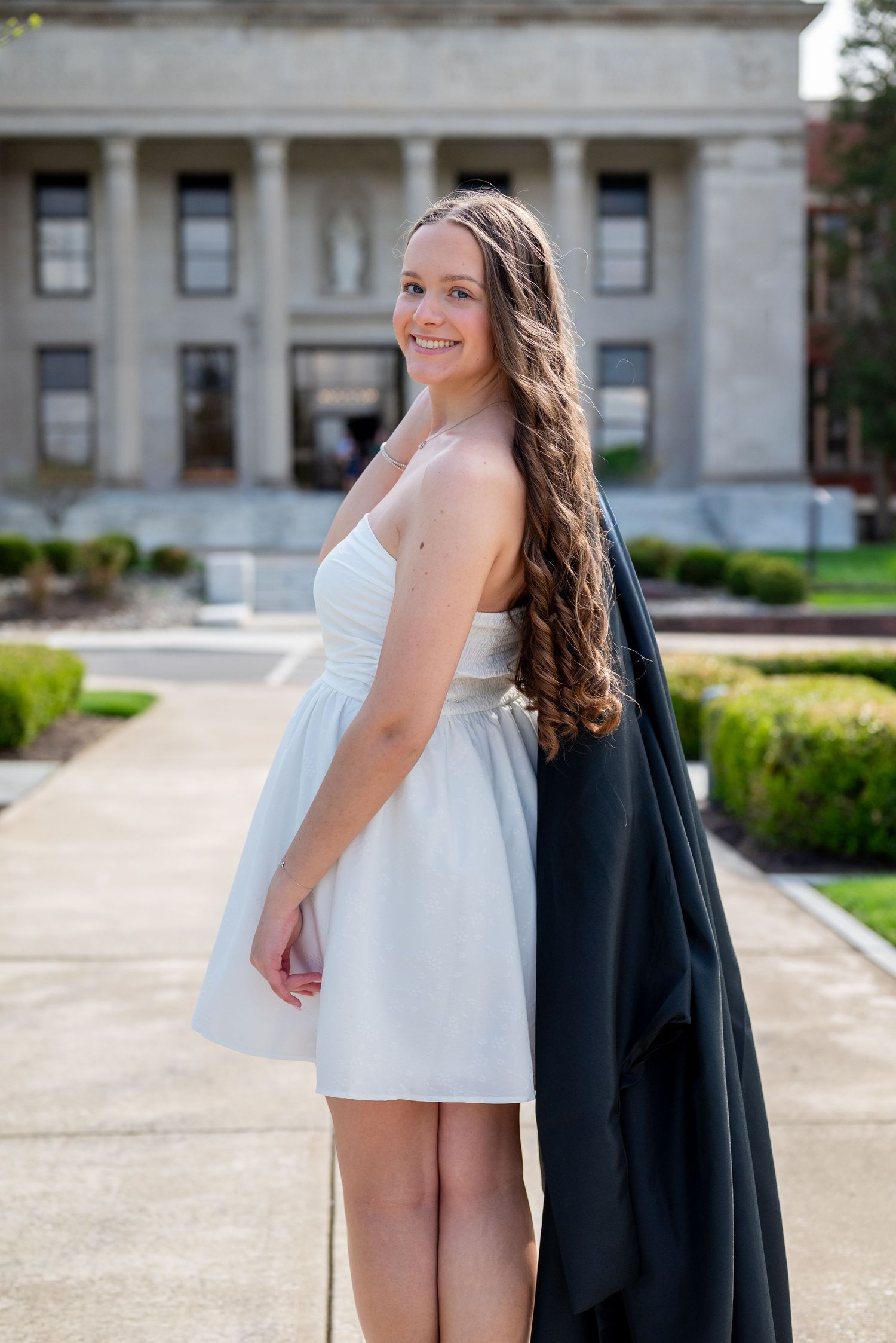 Smiling young woman in white dress holding black graduation gown outside a university building on a sunny day.