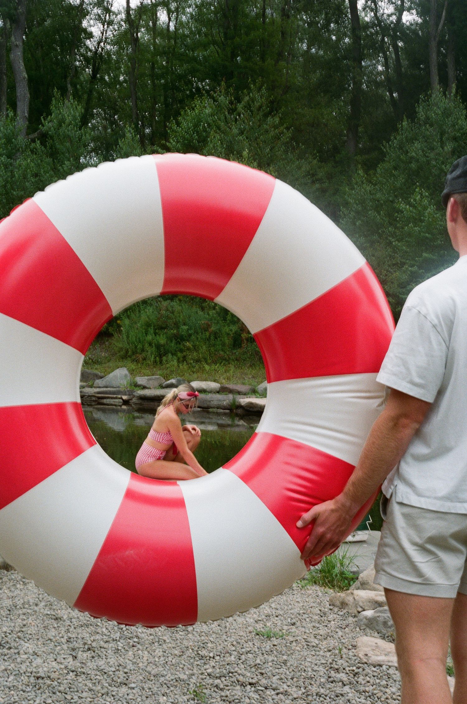 Man holds large red and white striped inflatable ring tube near a pond with a woman visible through the center hole.