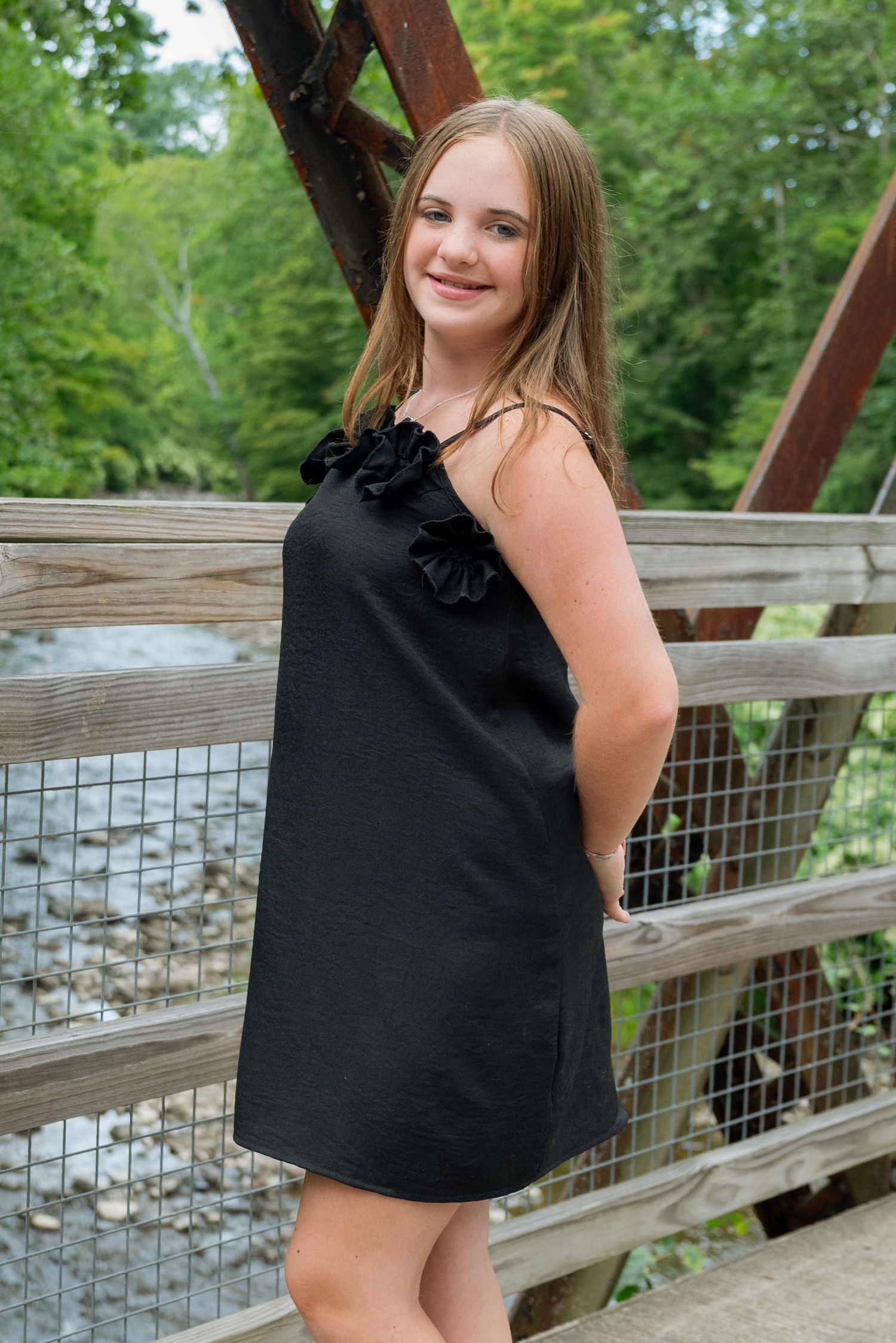 Young girl in black ruffle dress smiling on wooden bridge over stream surrounded by lush green trees.