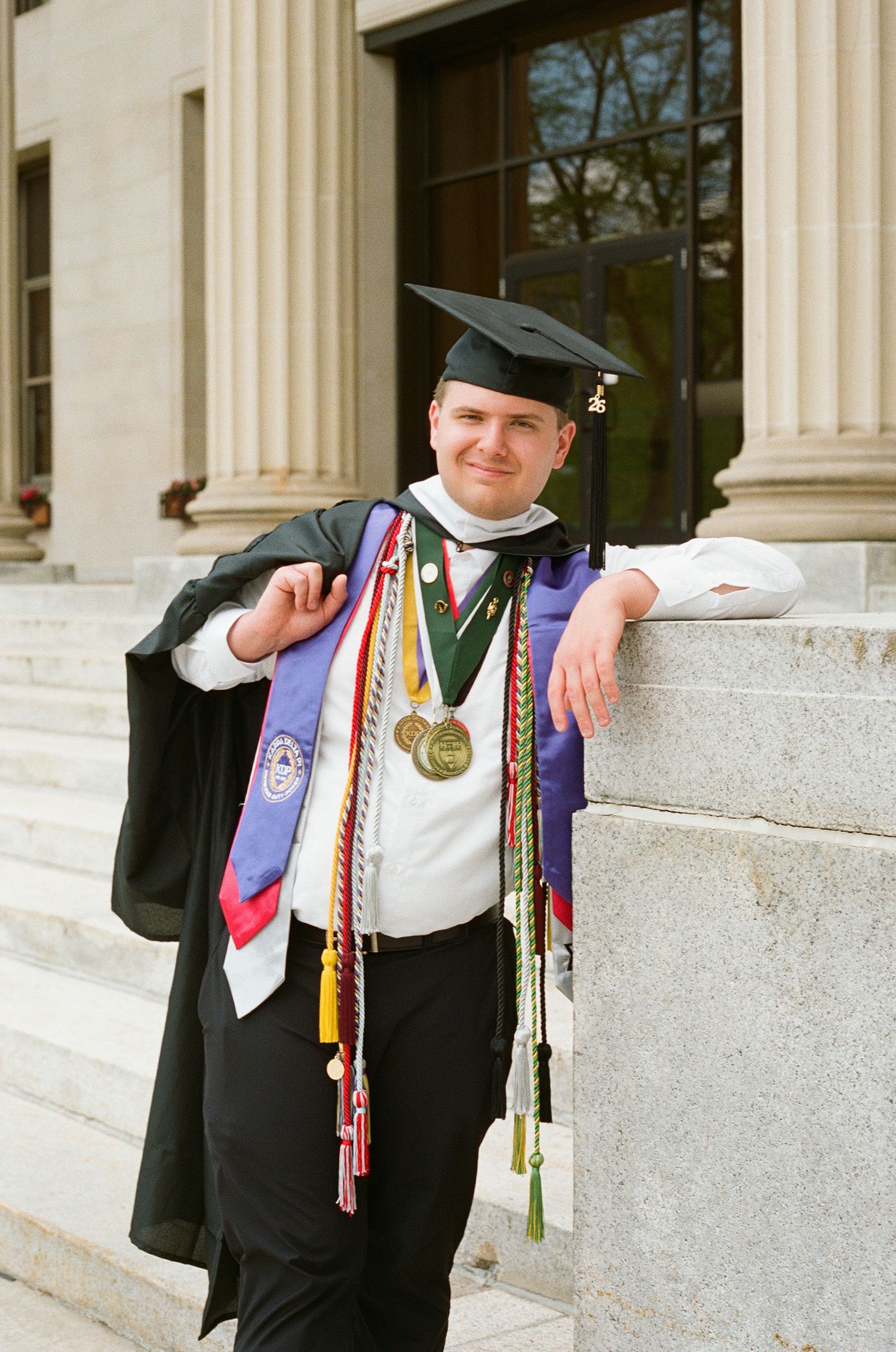 Graduate in cap and gown leaning on stone pillar wearing medals and honor cords at university steps.
