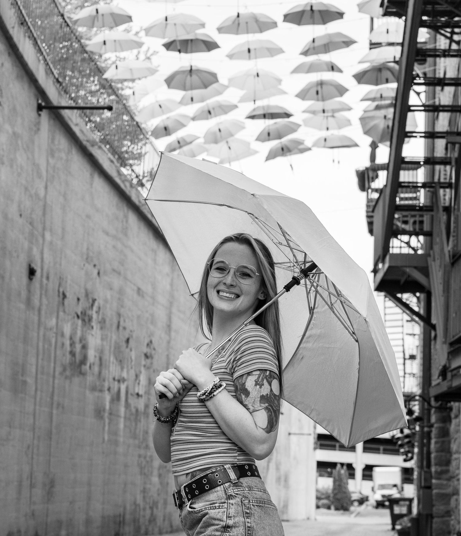 Smiling young woman holding umbrella under canopy of white umbrellas in urban alley, black and white photo.
