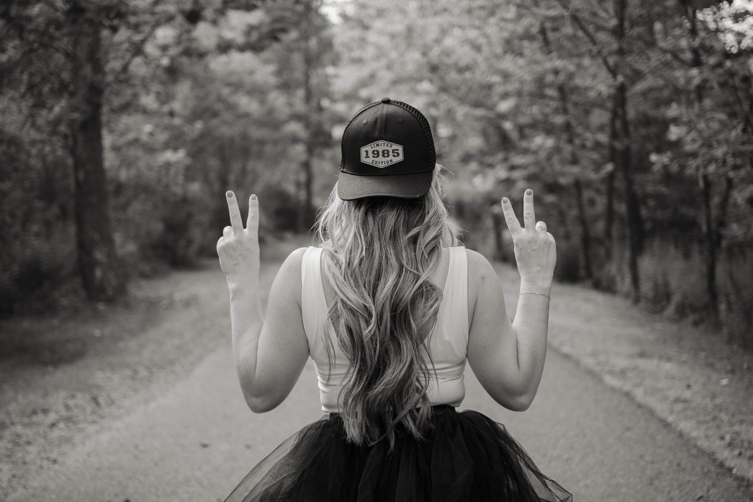 Woman in tulle skirt and cap makes peace signs on a tree-lined road in black and white photography.