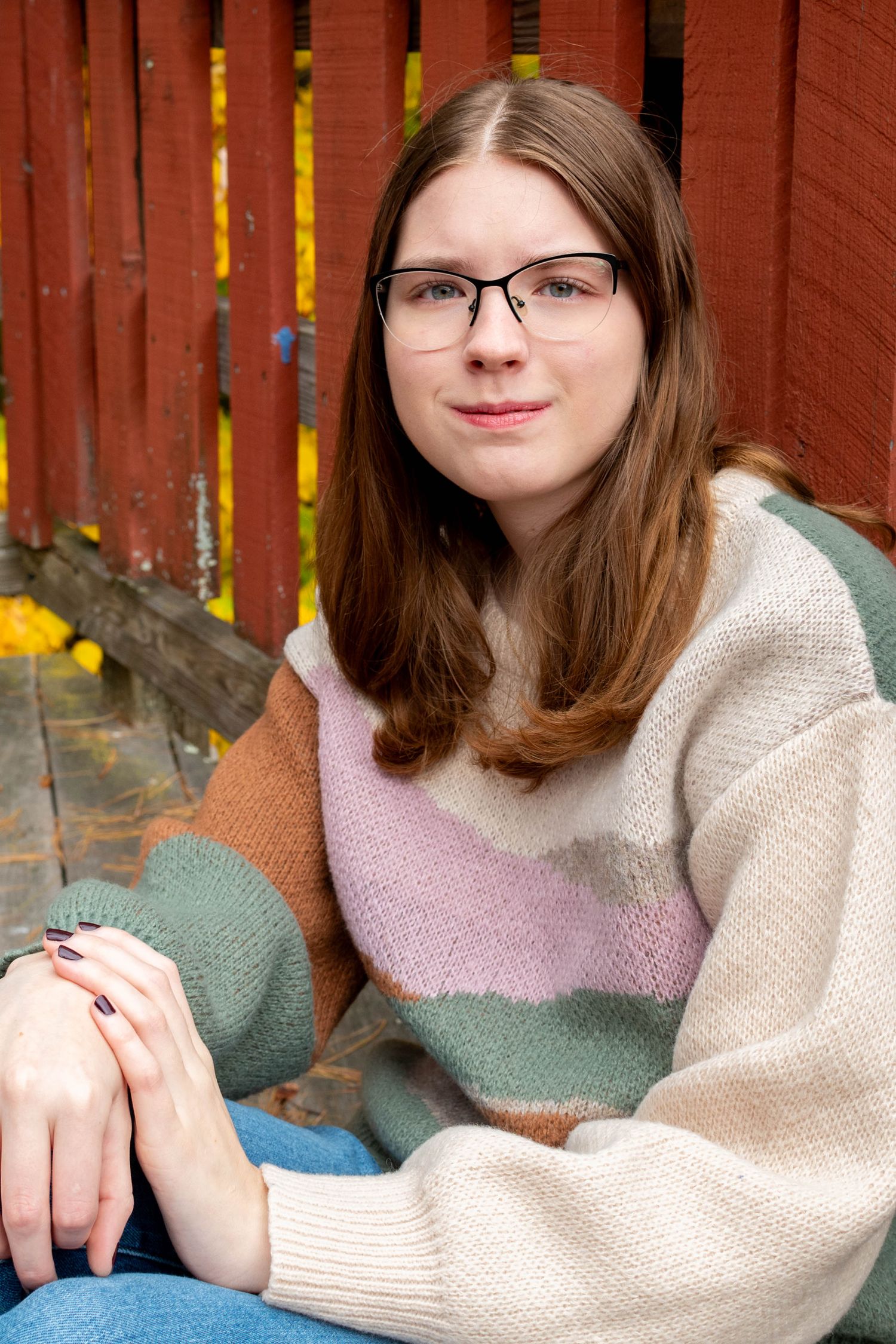 Teen girl with glasses in striped colorblock sweater sits relaxed near red wooden fence outdoors.