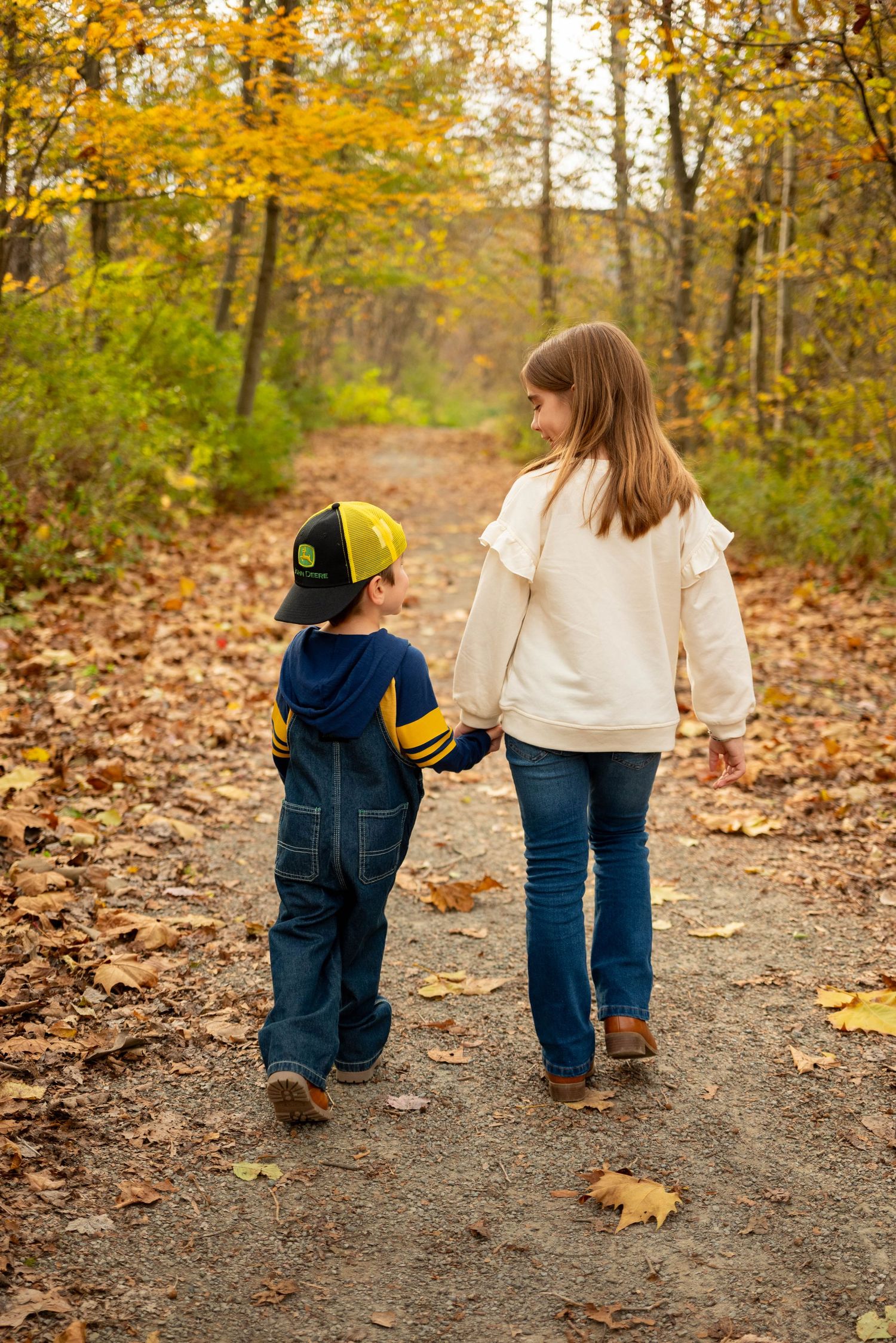 Two young siblings walk hand in hand down a leaf-covered autumn forest path from behind.