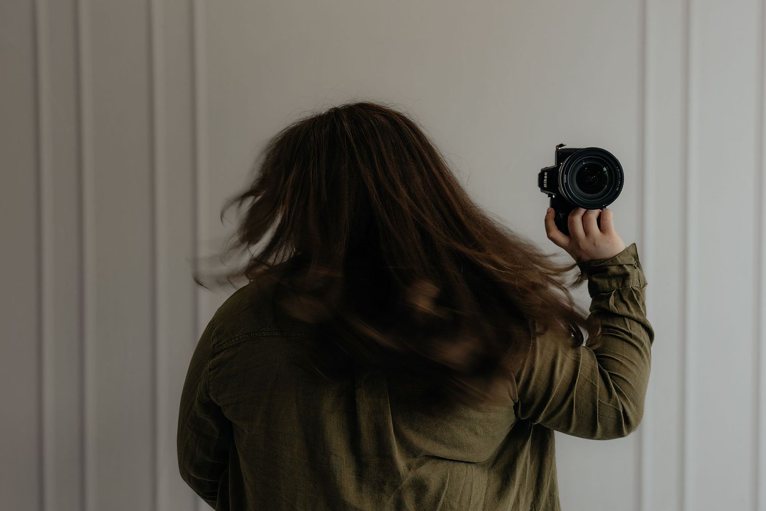 A person with long brown hair holds a camera, photographing a white wall, wearing an olive green jacket.
